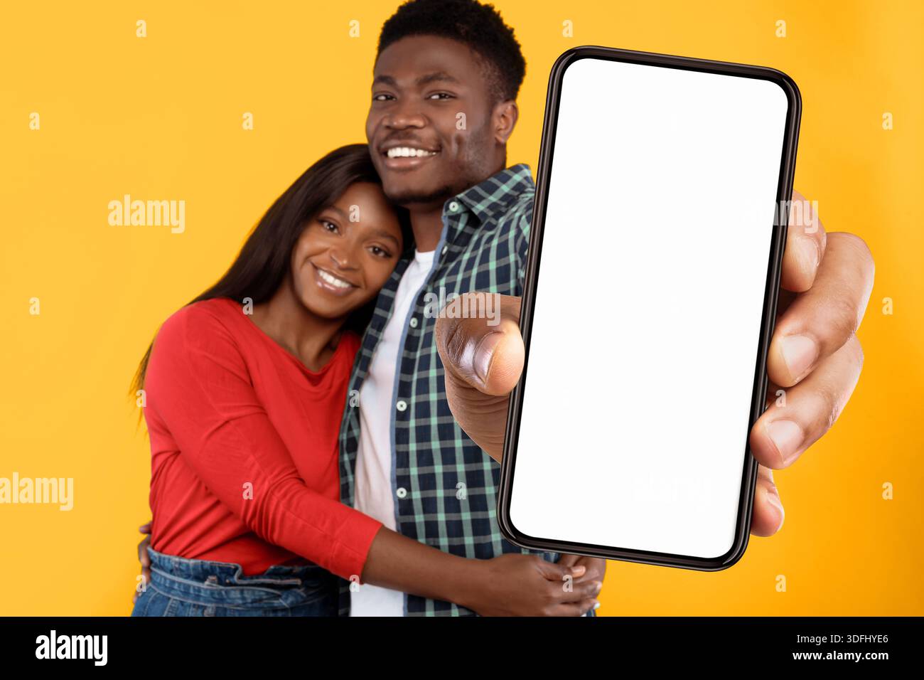 Ein Paar posiert zusammen und hält ein Smartphone vor einem leuchtend gelben Hintergrund Stockfoto