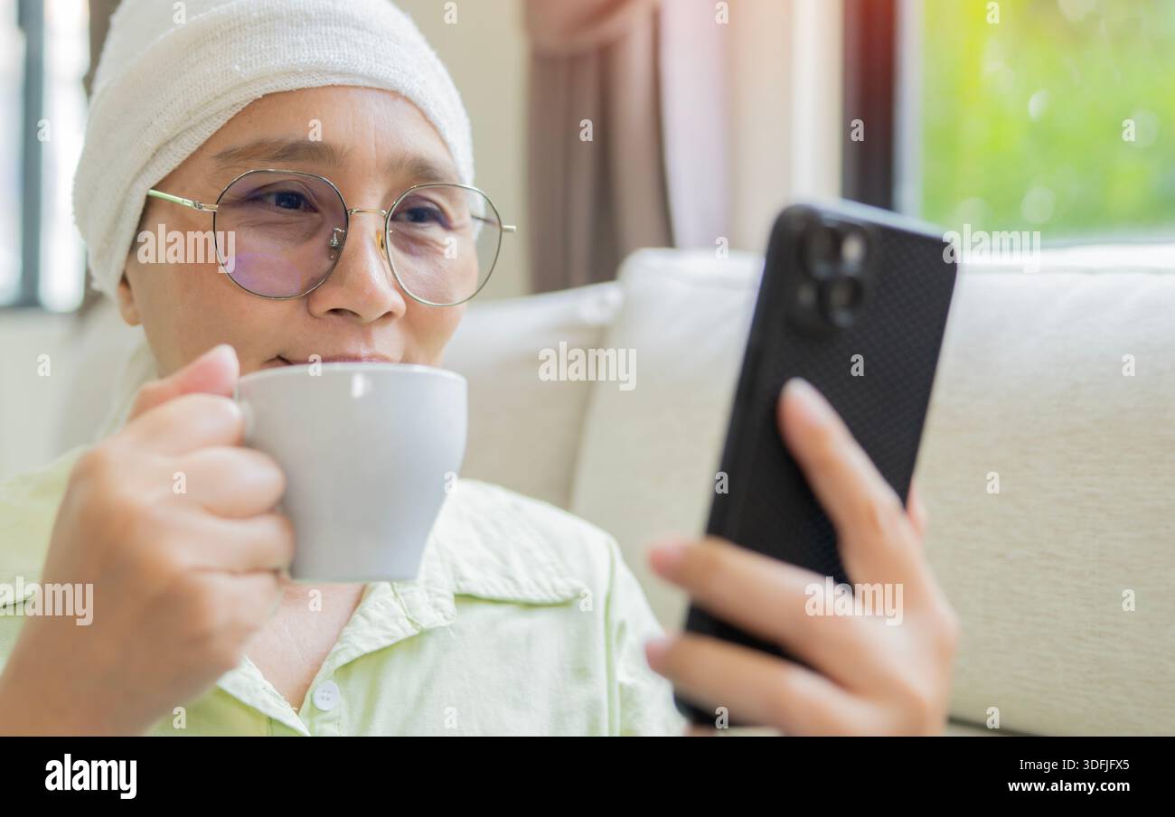 Frau mit Krebs und weißem Kopftuch, die auf das Handy blickt und neben dem Fenster lächelt, während sie eine Tasse Tee trinkt. Stockfoto