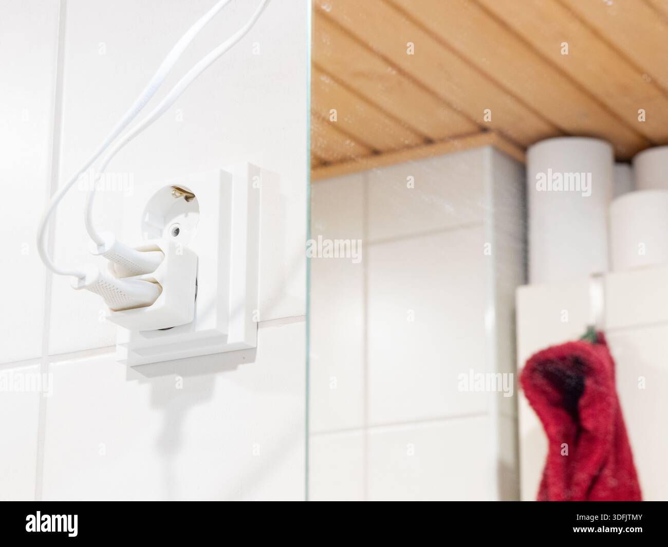 Eine weiße Steckdose mit Adapter verfügt über zwei Kabel, die an einer gekachelten Wand angeschlossen sind. Die rechte Seite zeigt eine Spiegelreflektion der Holzdecke, t Stockfoto