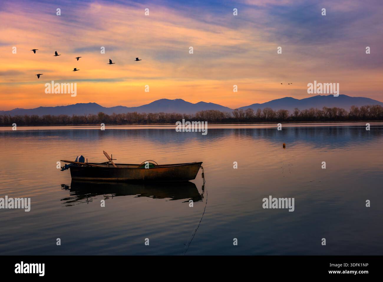 Kerkini, Nordgriechenland – Ein hölzernes Fischerboot ruht auf ruhigen Gewässern bei Sonnenuntergang, während Vögel in einer ruhigen, friedlichen Szene über den glühenden Himmel fliegen. Stockfoto