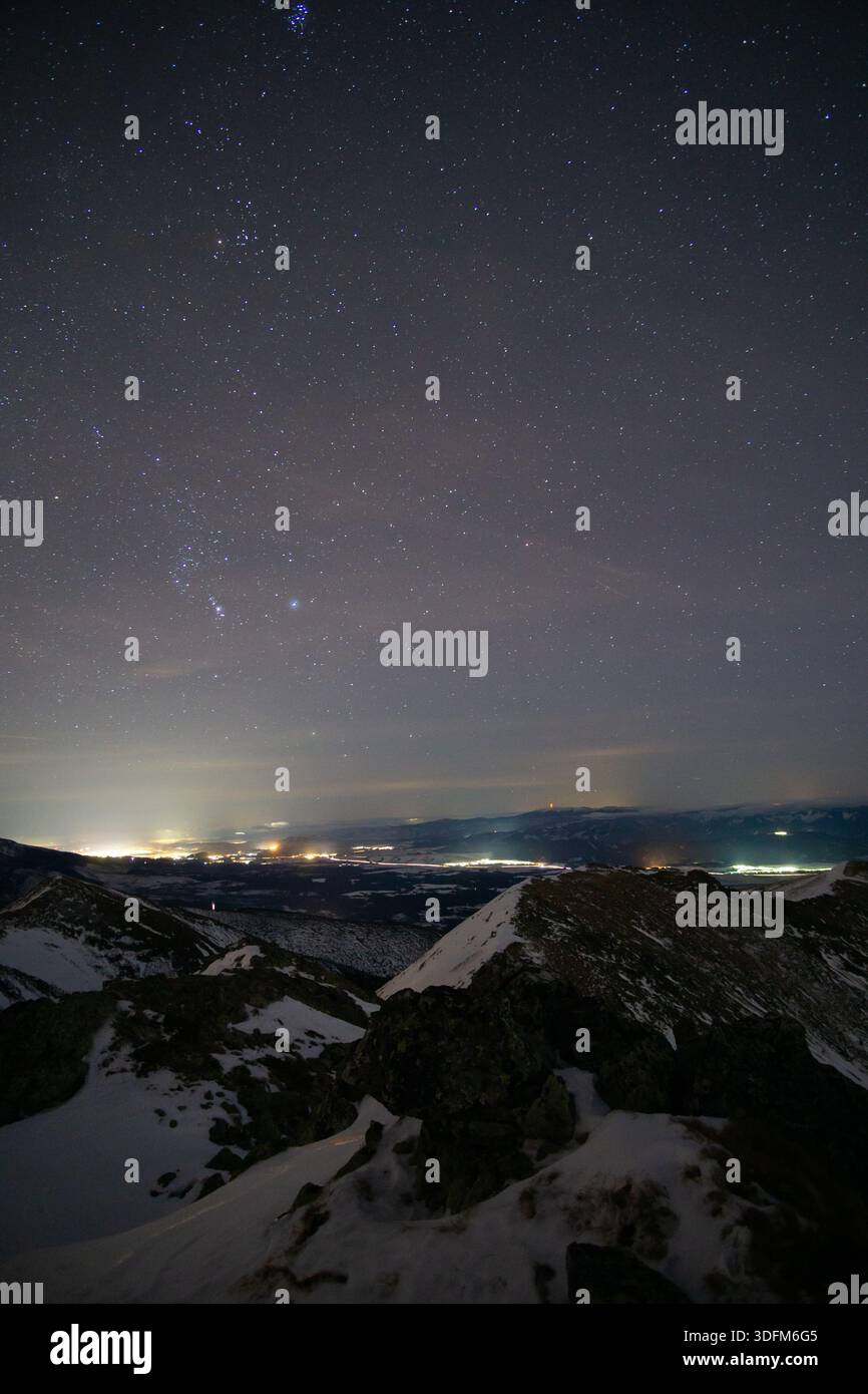 Blick auf die funkelnden Sterne über den schneebedeckten Gipfeln, die ein heiteres Leuchten über die fernen Lichter der Stadt ausstrahlen, eine ruhige Szene, Pribylina, Zilina Regio Stockfoto