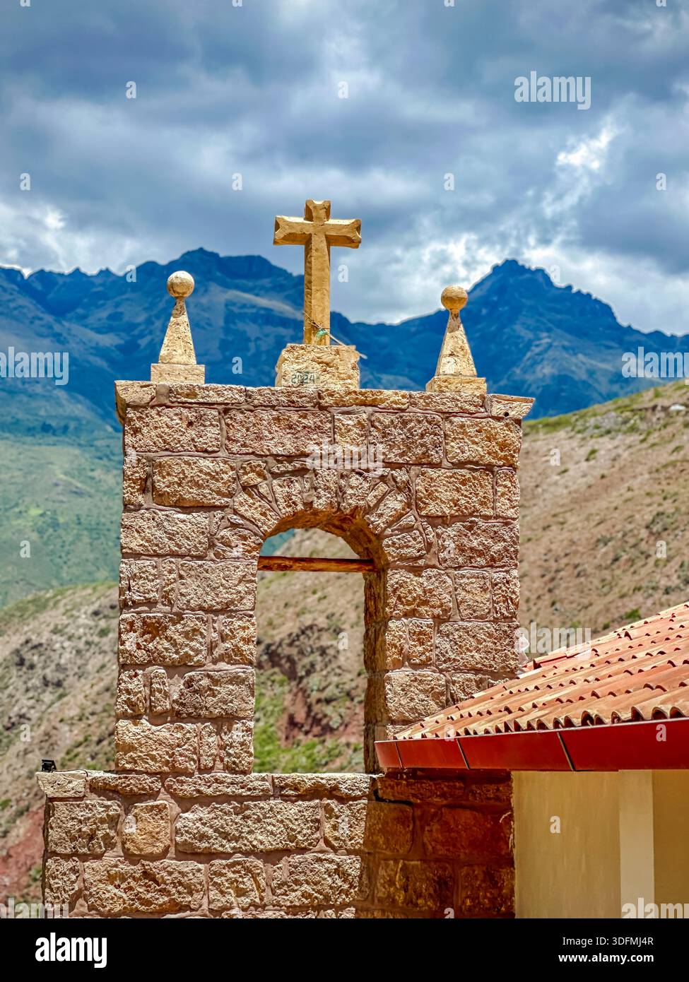 Blick auf einen rustikalen Steinbogen mit einem goldenen Kreuz vor der Kulisse majestätischer Berge und dramatischer Himmel, Maras, Peru. Stockfoto