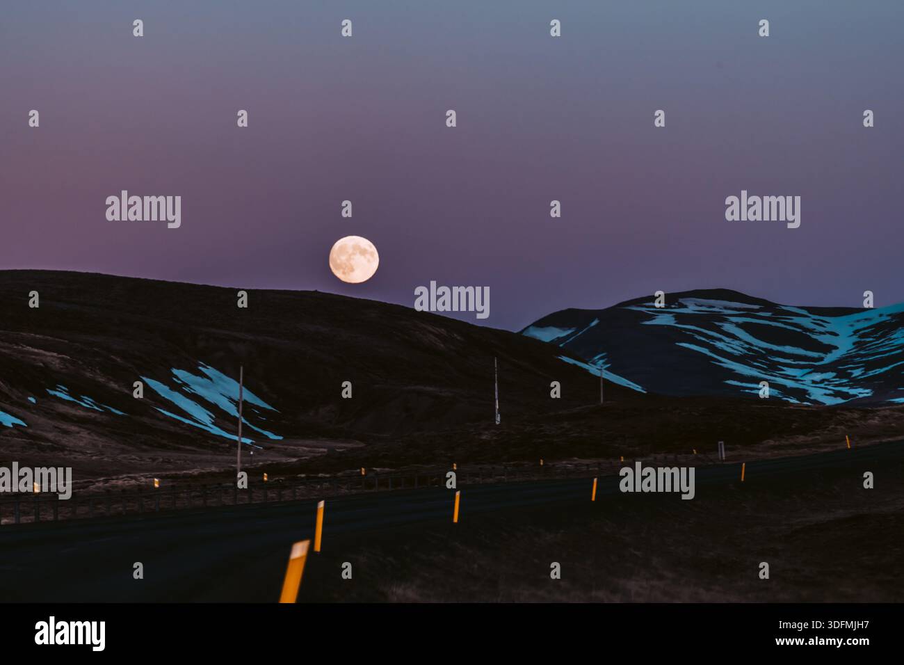 Der Blick auf einen faszinierenden Vollmond hängt in der Abenddämmerung über dunklen, sanften, von Schnee berührten Hügeln, über einer von gelben Markierungen gesäumten steilen Straße, Saudar Stockfoto