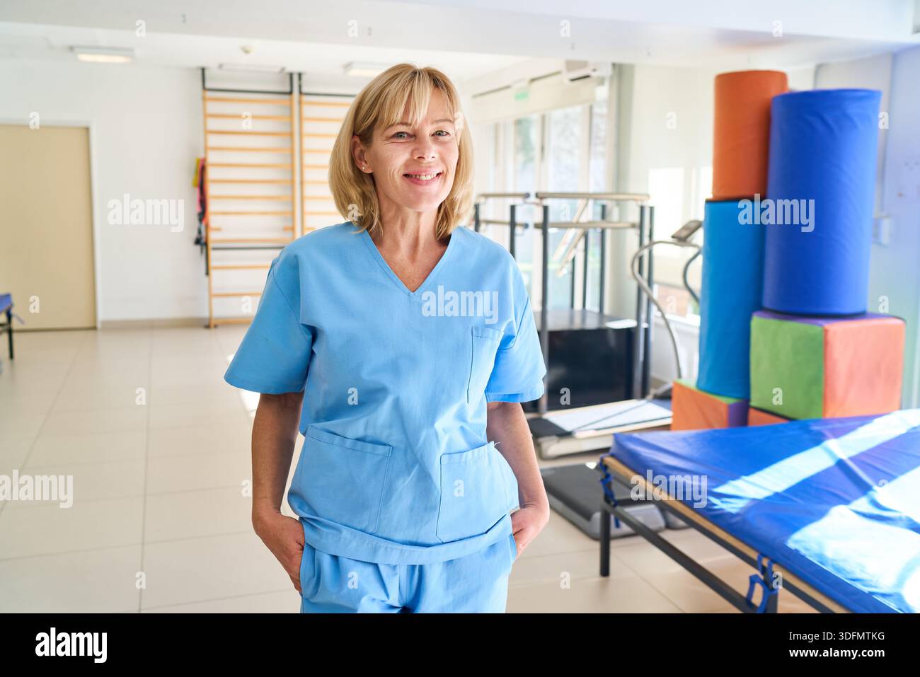 Eine lächelnde reife Frau in blauen Schutten steht in einem Rehabilitationszentrum mit Trainingsgeräten und einem Arzt. Diese Abbildung dient zur Veranschaulichung Stockfoto