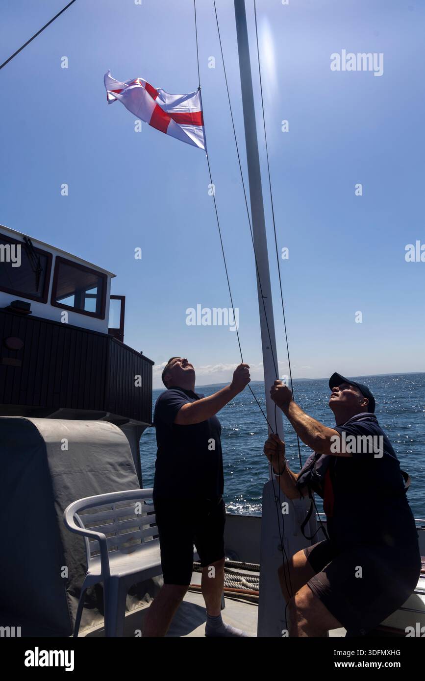 Die Besatzung der HMT hisst die ADLS-Flagge von St. George, während das Schiff wieder in britische Gewässer einfährt. Stockfoto