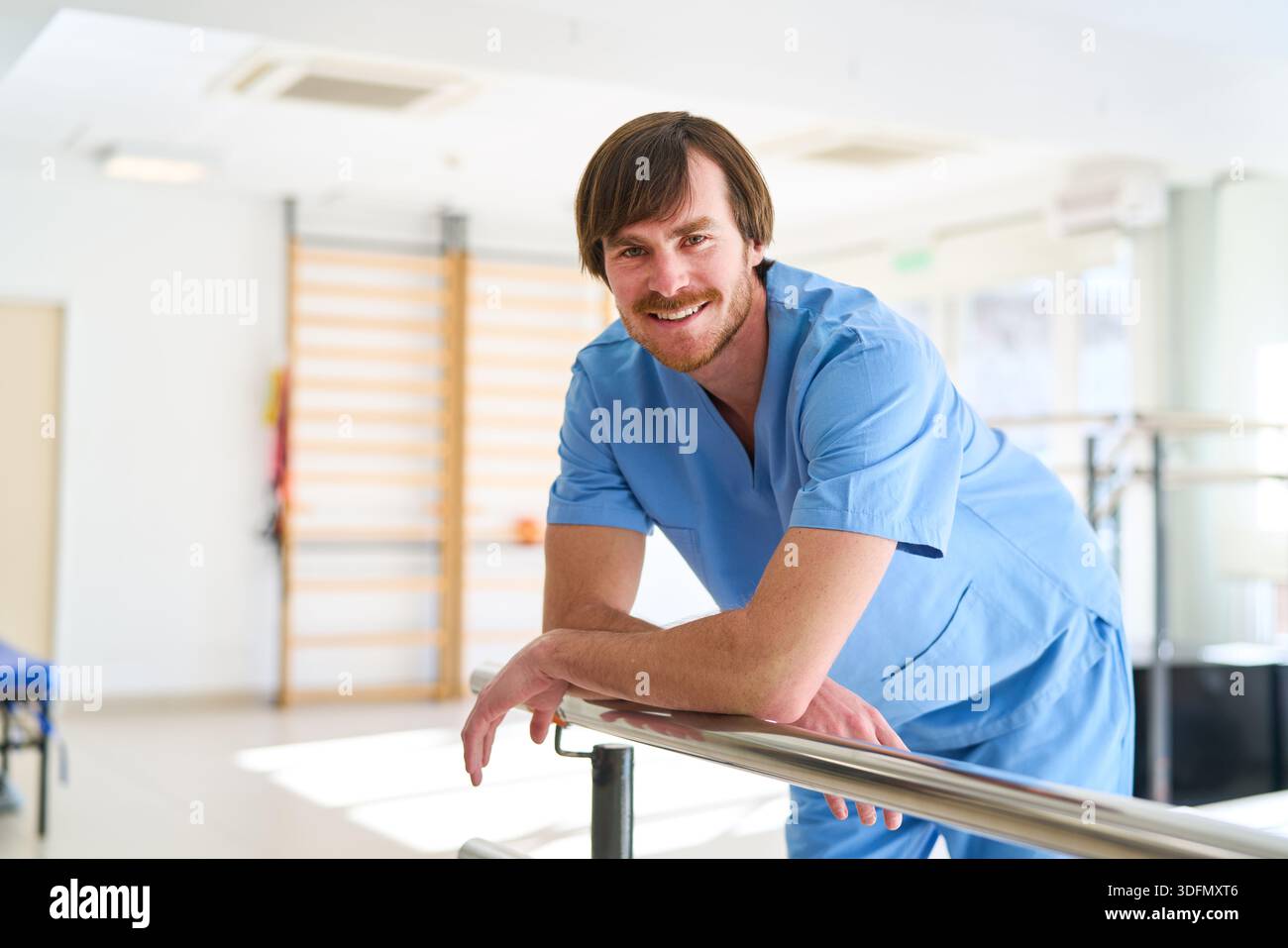 Ein lächelnder Physiotherapeut in blauem Scrubs beugt sich an parallelen Gittern in einem Rehabilitationszentrum. Diese Abbildung ist nützlich, um die Gesundheitsfürsorge darzustellen Stockfoto
