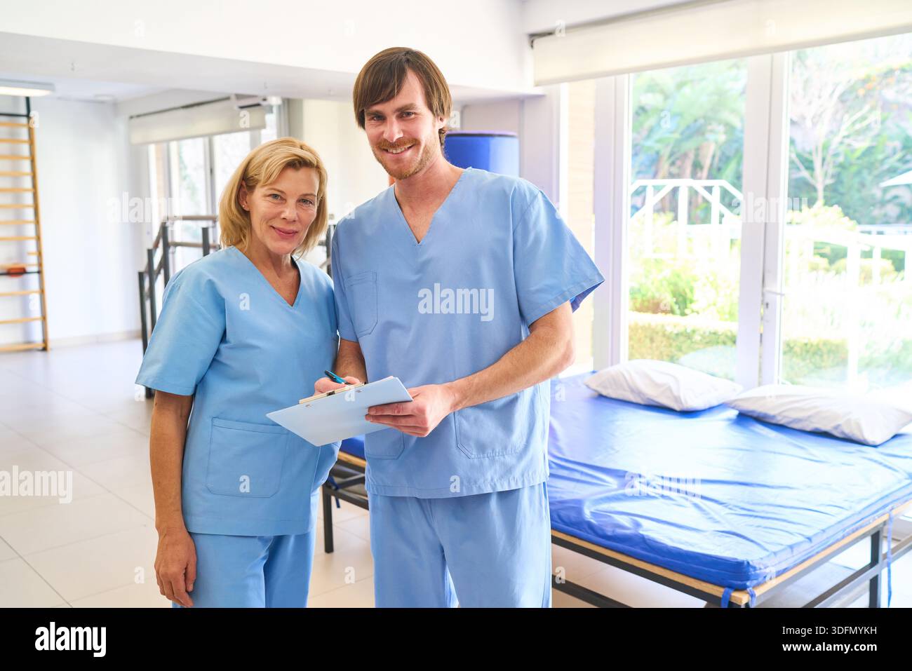 Zwei Physiotherapeuten in blauen Peelings stehen in einem Reha-Zentrum, einer hält ein Klemmbrett, das einem älteren Patienten Erholung und Rehabilitation in Innenräumen bietet. Stockfoto