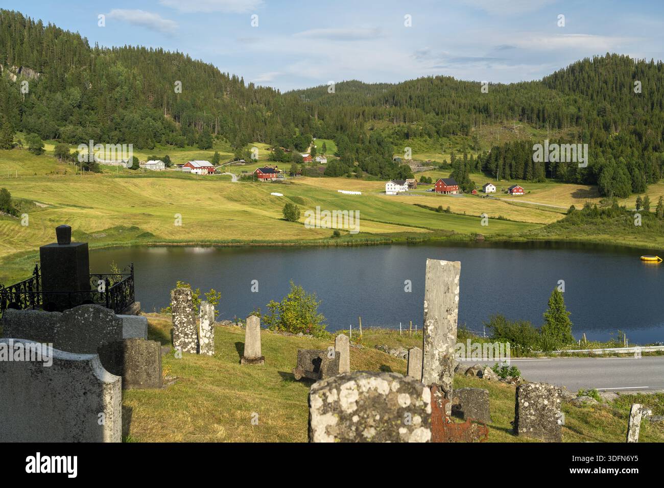 Blick auf einen ruhigen See, der den Himmel spiegelt, eingebettet in sanfte Hügel, mit einem historischen Friedhof im Vordergrund, der einen ergreifenden Kontrast bildet: Eidsborg, Stockfoto
