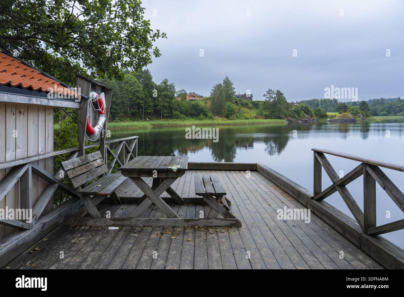 Blick auf den ruhigen See, der den Himmel reflektiert, von einer Holzterrasse mit Bänken und einem an einer Wand hängenden Rettungsschwimmer, Treungen, Telemark, Norwegen. Stockfoto