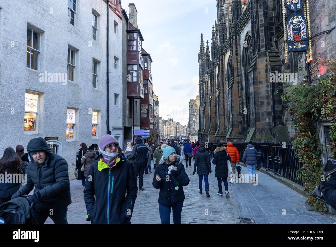 Eine geschäftige Straßenszene, in der Fußgänger in Winterkleidung an historischen Gebäuden vorbeilaufen, darunter eine gotische Kirche und Geschäfte in Edinbur Stockfoto