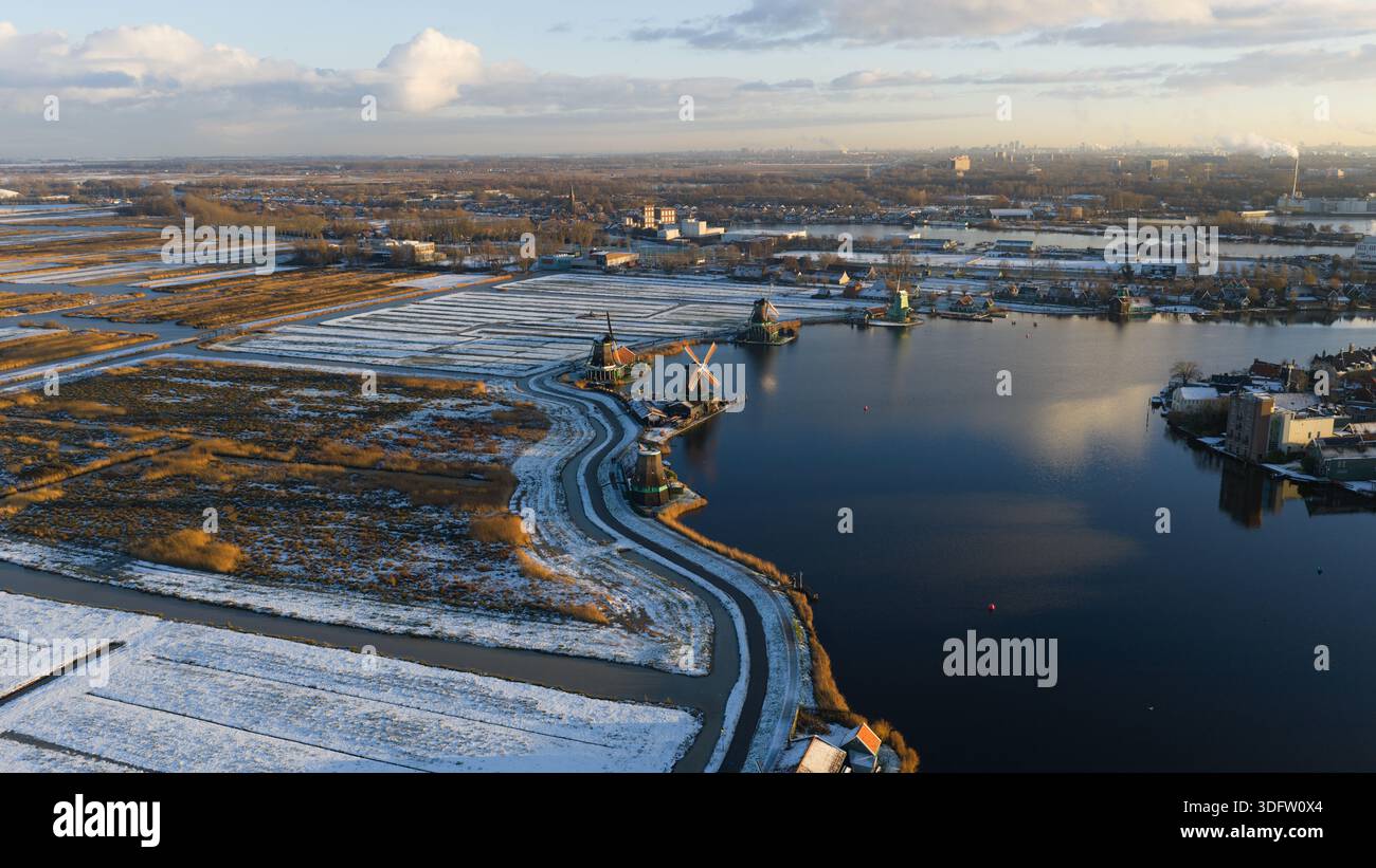 Aus der Vogelperspektive von Windmühlen, die stolz gegen das kalte Wasser stehen, eingerahmt von schneebedeckten Feldern unter einem weichen Winterhimmel, Zaanse Schans, Niederlande. Stockfoto