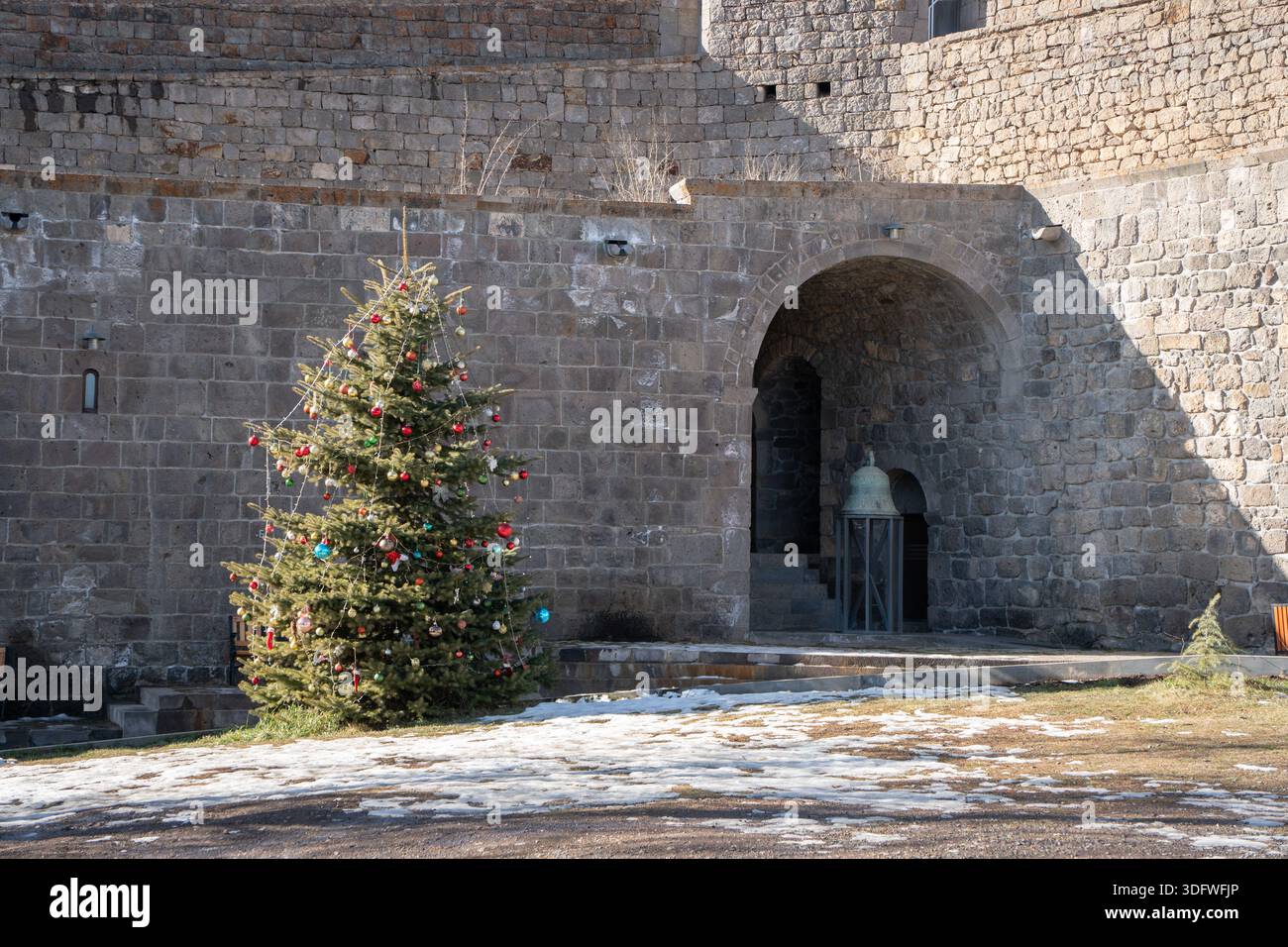 Tatev, Armenien - 08. Januar 2026: Geschmückte Tannenbäume stehen im Innenhof in der Nähe eines Steinbogeneingangs, befestigte Klostermauer, Schneebesen Stockfoto