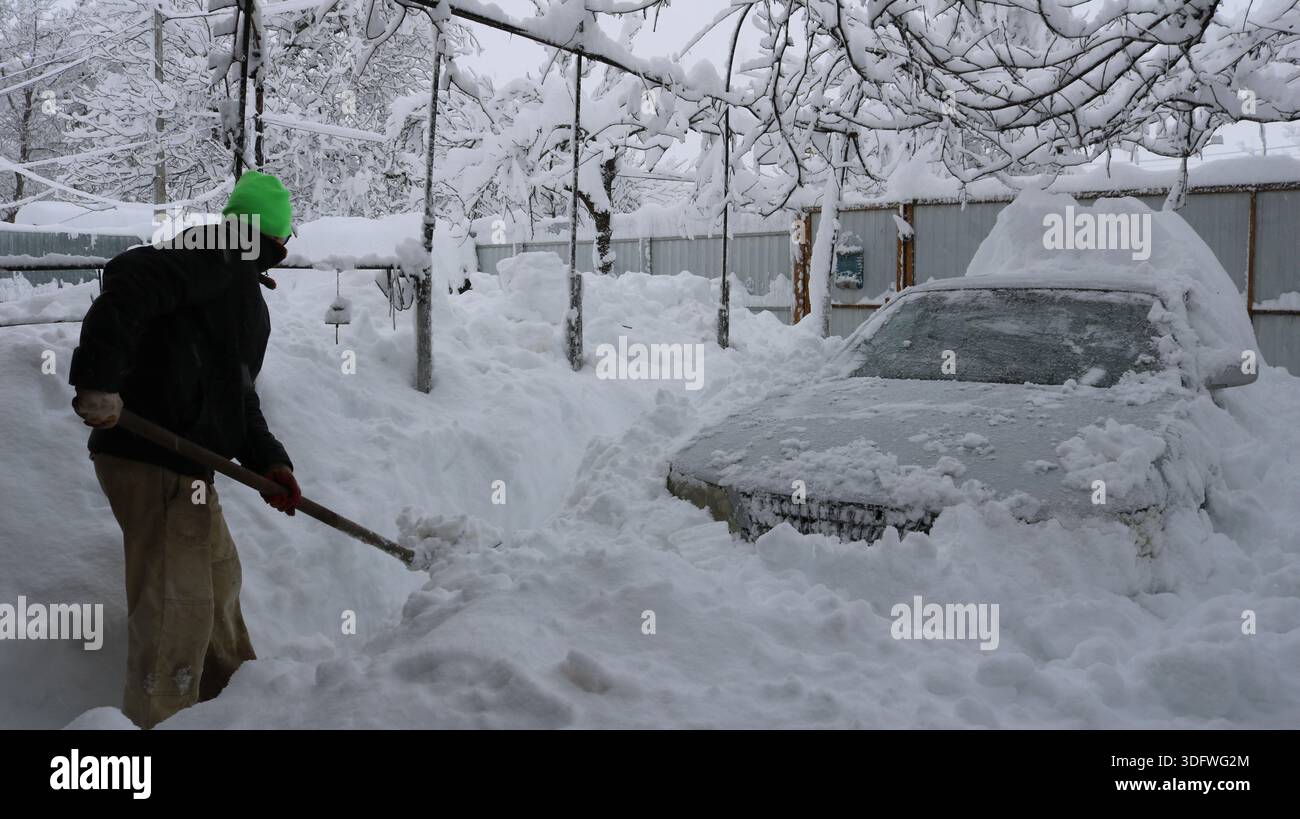 Ein Mann in Winterkleidung räumt mit einer Holzschaufel den Weg zu einem schneebedeckten Auto in einem ländlichen Hof und gräbt manuell ein graues Auto aus Stockfoto