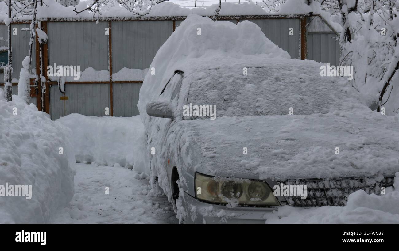 Ein graues Auto, teilweise von Schnee befreit auf einem privaten Hof unter einer leichten Frostschicht, parkt zwischen tiefen Schneelagen nach ungewöhnlichem Wetter Stockfoto