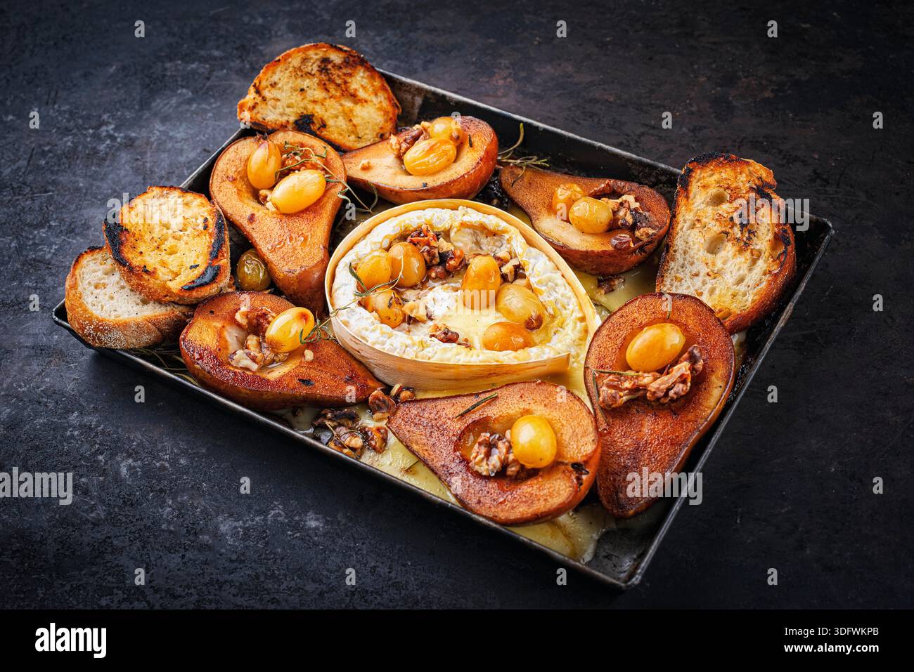 Im Ofen gebackener Camembert-Käse mit karamelisierten glasierten Birnen, Walnüssen und Trauben sowie Baguette auf rustikalem Metallteller Stockfoto