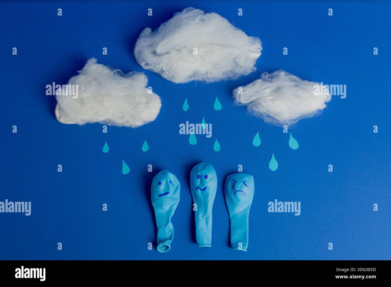 Drei blaue Luftballons mit traurigen Gesichtern unter Baumwollwolken und Regentropfen auf blauem Hintergrund. Stockfoto