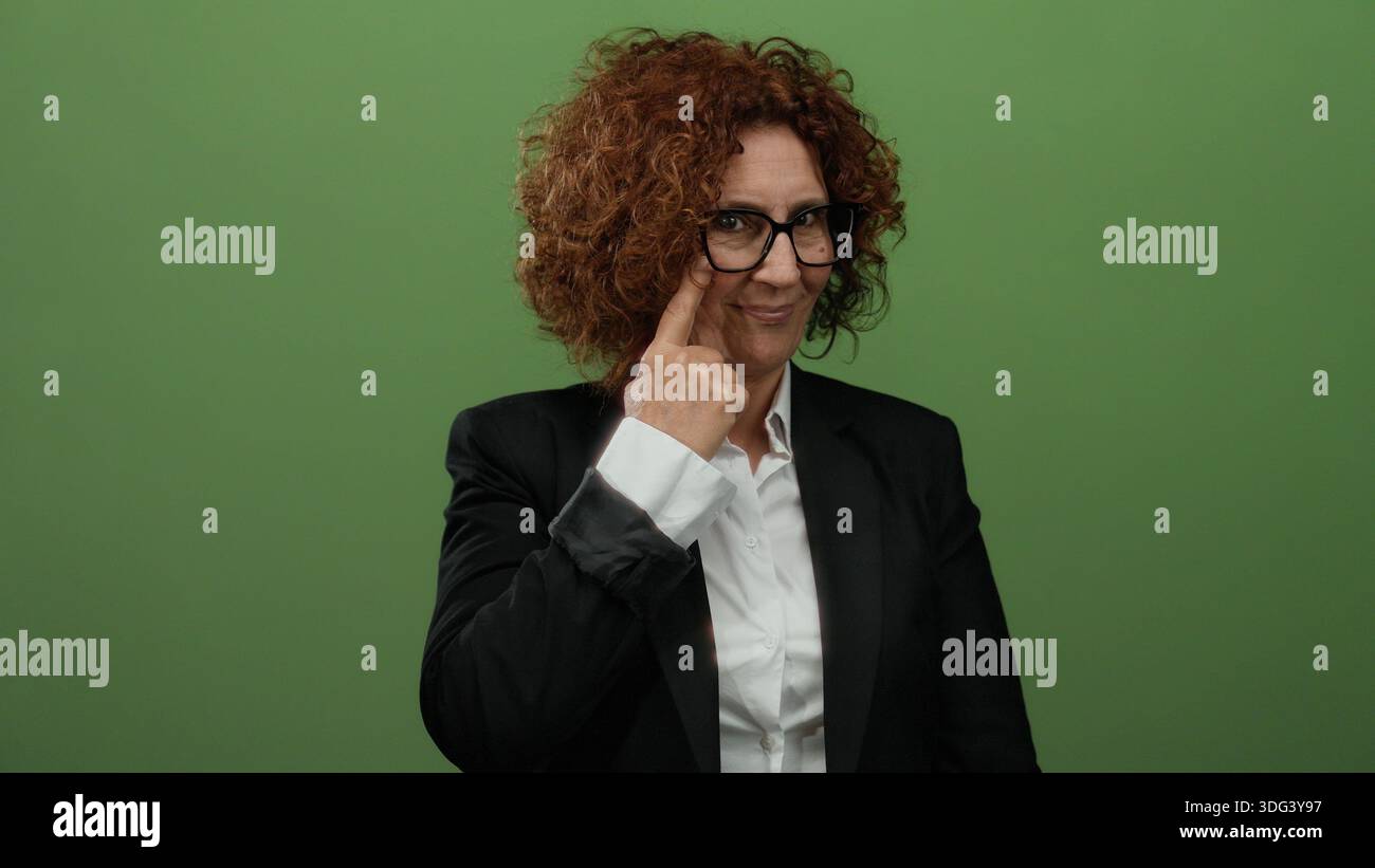 Frau mit Brille, die eine wachsame Geste macht, steht vor einem grünen Hintergrund, trägt einen formellen Anzug, zeigt ein Gefühl von Bewusstsein und Stockfoto
