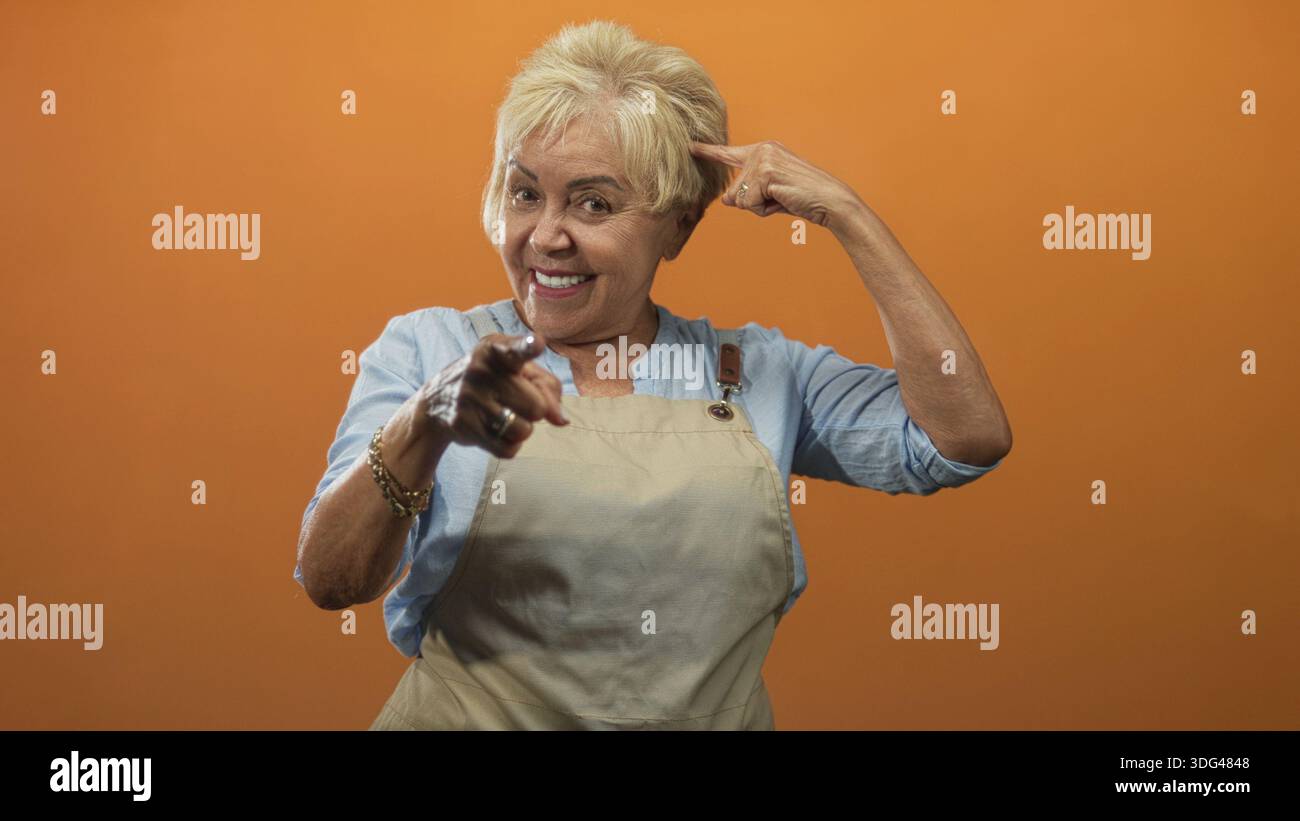 Die ranghöchste hispanische Frau in der Schürze zeigt den Finger auf die Kamera und zeigt den Finger zum Tempel in einem Studio mit orangefarbenem Hintergrund; spielerisches Selbstvertrauen. Stockfoto