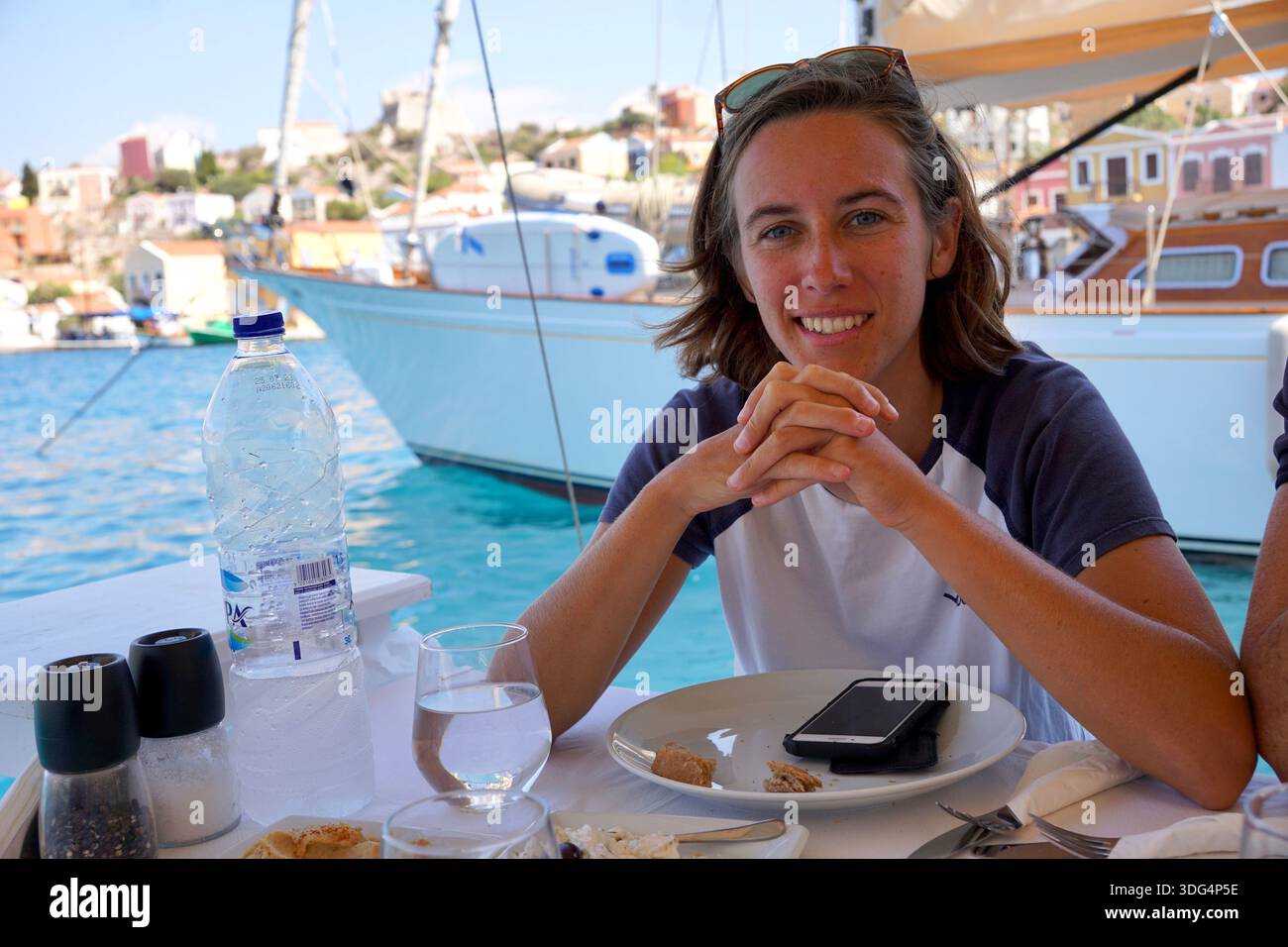 Junge Frau, die am Tisch im Freien am Hafen auf der griechischen Insel Kastellorizo sitzt und mit Yachten und türkisfarbenem Wasser dahinter lächelt. Stockfoto