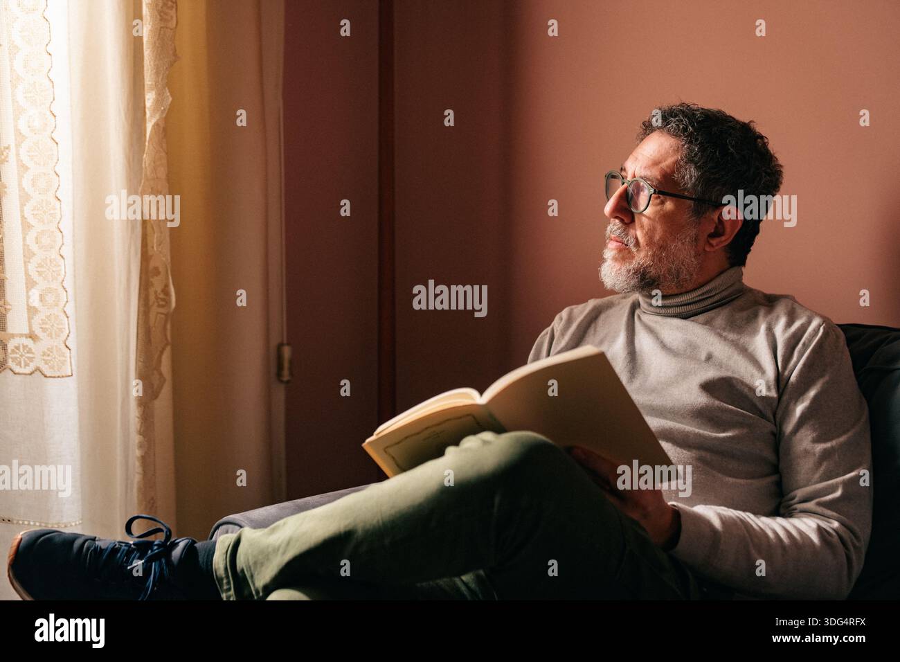 Mann mittleren Alters, der auf dem Sofa sitzt und ein Buch hält, während er aus dem Fenster blickt, in Gedanken verloren. Ruhiger Moment im Innenbereich, der Besinnung und langsames Leben ausdrückt Stockfoto
