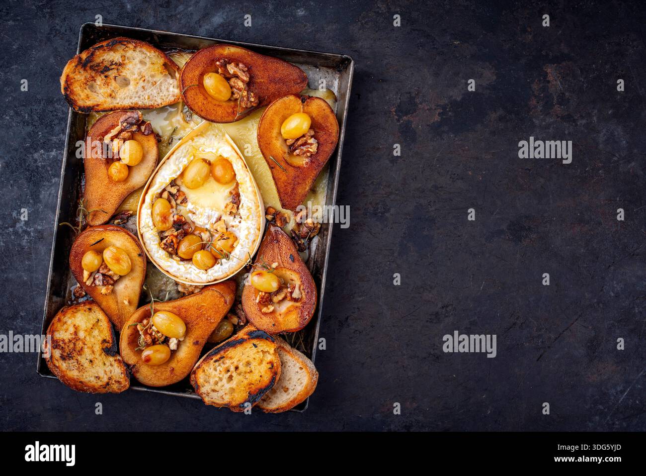 Im Ofen gebackener Camembert-Käse mit karamelisierten glasierten Birnen, Walnüssen und Trauben sowie Baguette auf rustikalem Metallteller Stockfoto