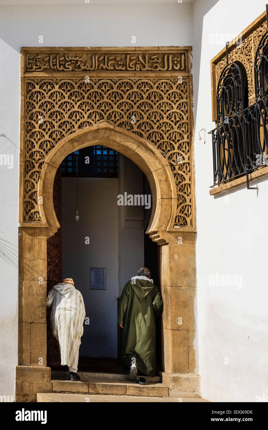 Zwei Personen in traditioneller Kleidung betreten einen kunstvollen bogenförmigen Eingang mit komplizierten Schnitzereien. Rabat, Marokko Stockfoto
