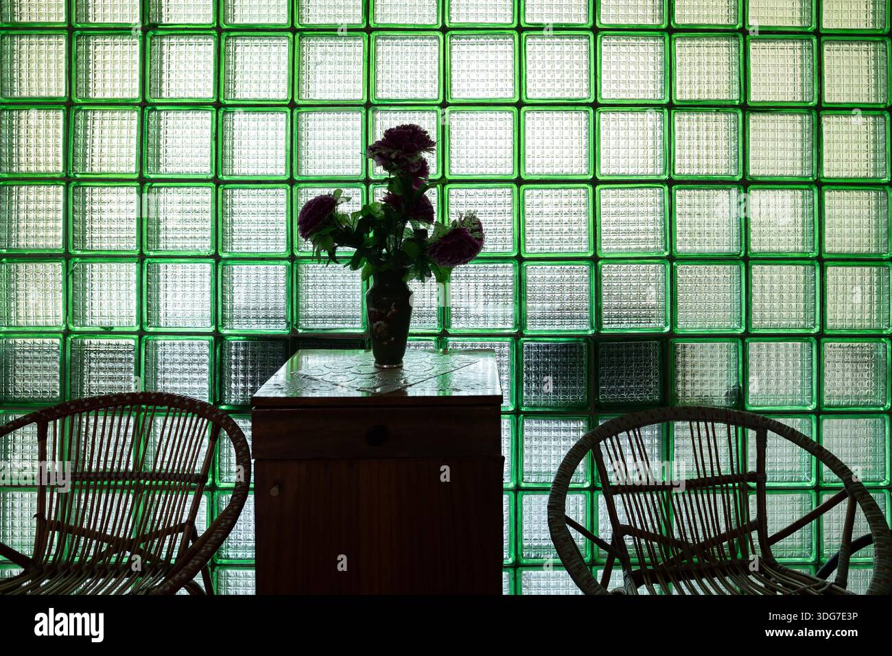 Retro-Inneneinrichtung mit einer Trennwand aus grünem Glasblock mit einer Blumenvase auf einem Schrank und Korbstühlen. Nostalgische Ästhetik und Deko Stockfoto