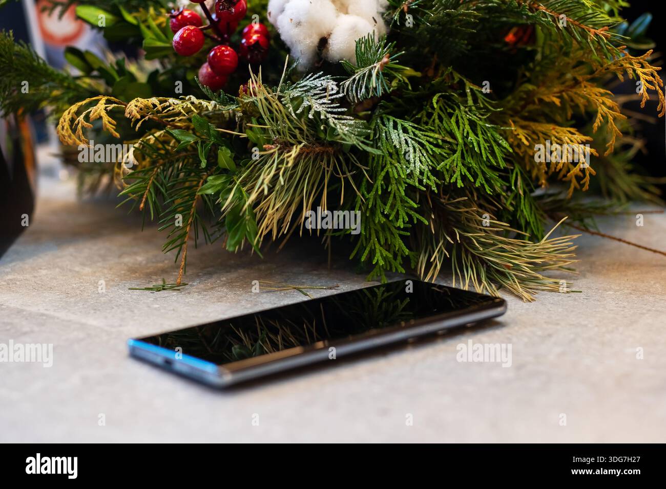 Verlorenes Smartphone unter festlichem, immergrünem Herzstück, glänzendes schwarzes Gerät, das auf Steintisch neben holly Beeren liegt Stockfoto