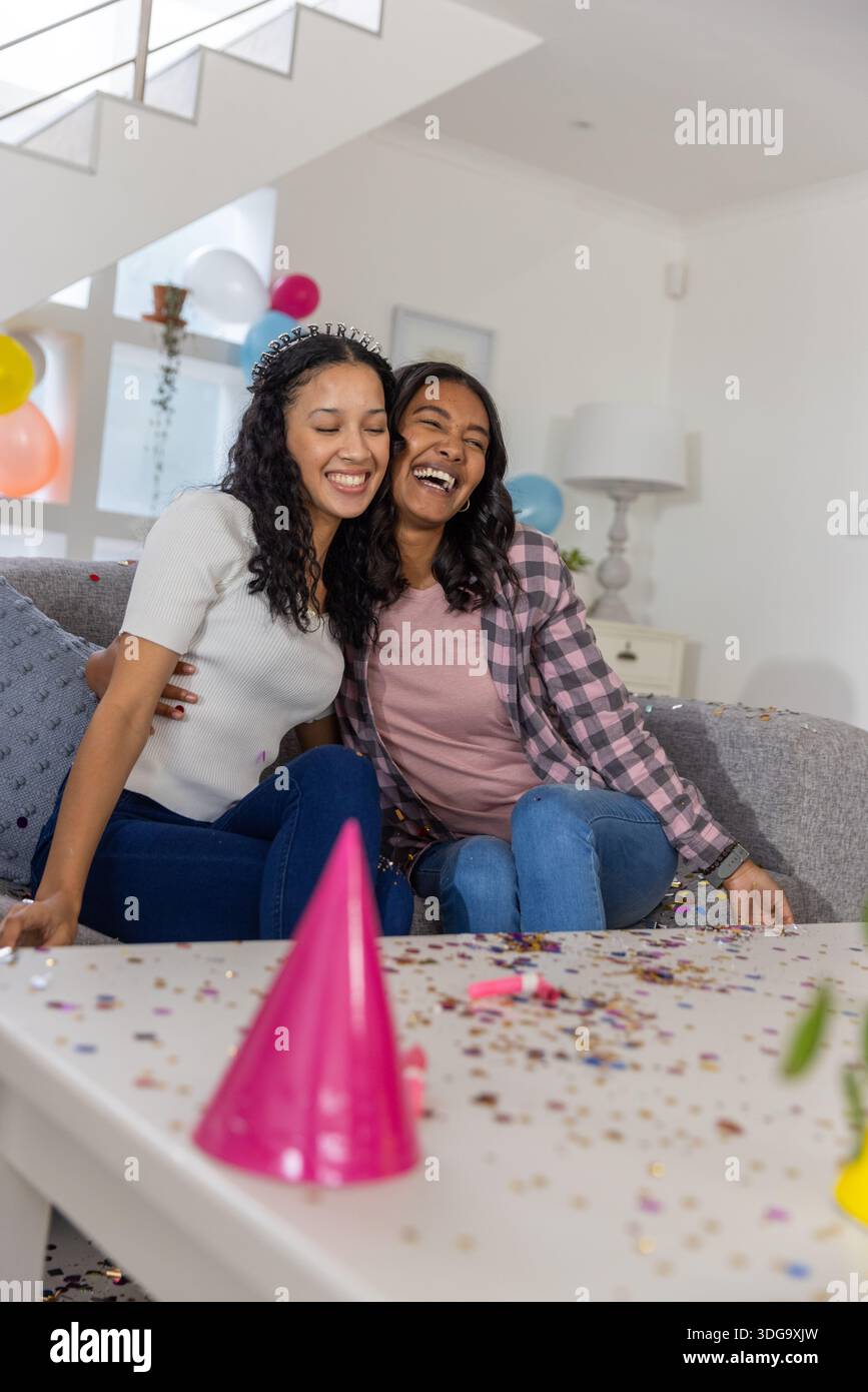 Vielfältige Mutter und Teenager-Tochter umarmen und lachen auf dem Sofa im Wohnzimmer mit Ballons Stockfoto