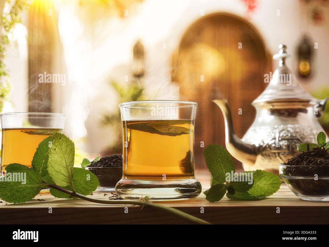 Details zum Gießen marokkanischer Teekanne aus Metall in ein Glas auf einem Holztisch mit einer Schüssel getrockneten Blatttees, dekoriert mit Minzblättern nach einer Tradition Stockfoto