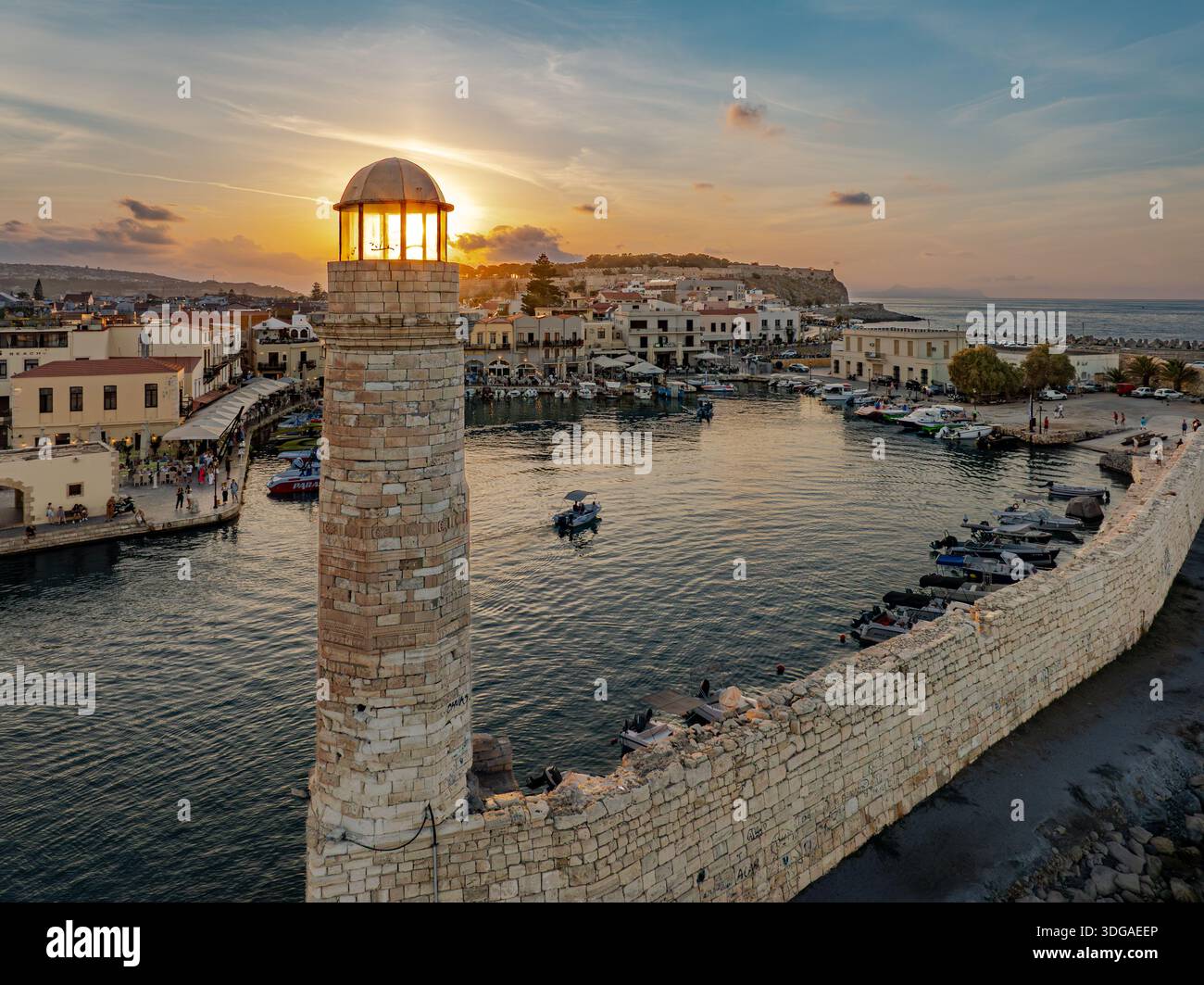 Aus der Vogelperspektive auf den sonnengeküssten Leuchtturm, der über dem ruhigen Hafen mit Booten wacht, eingerahmt von den alten venezianischen Mauern Rethimno, Re Stockfoto