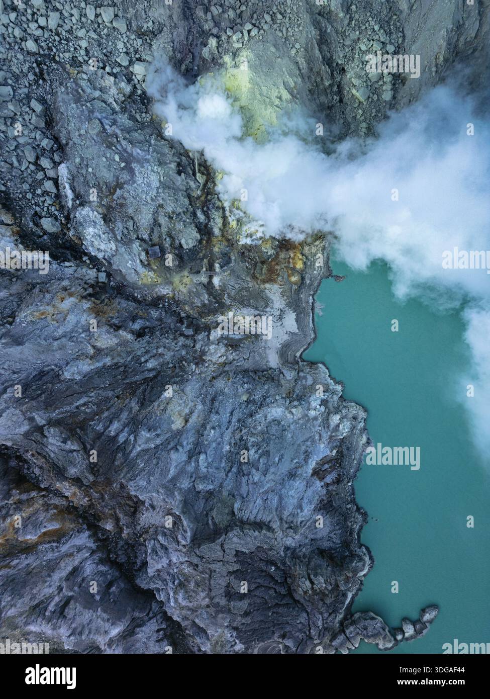 Aus der Vogelperspektive auf den türkisfarbenen See, der die strukturierte, felsige Landschaft trifft, mit weißen Rauchschwaden, die in die Atmosphäre aufsteigen, Banyuwangi, Ost-Java, Stockfoto