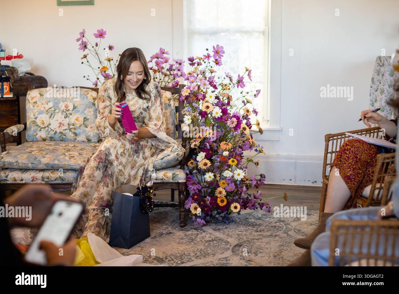 Frau auf Blumensofa packt Babyduschgeschenke aus, umgeben von Fl Stockfoto