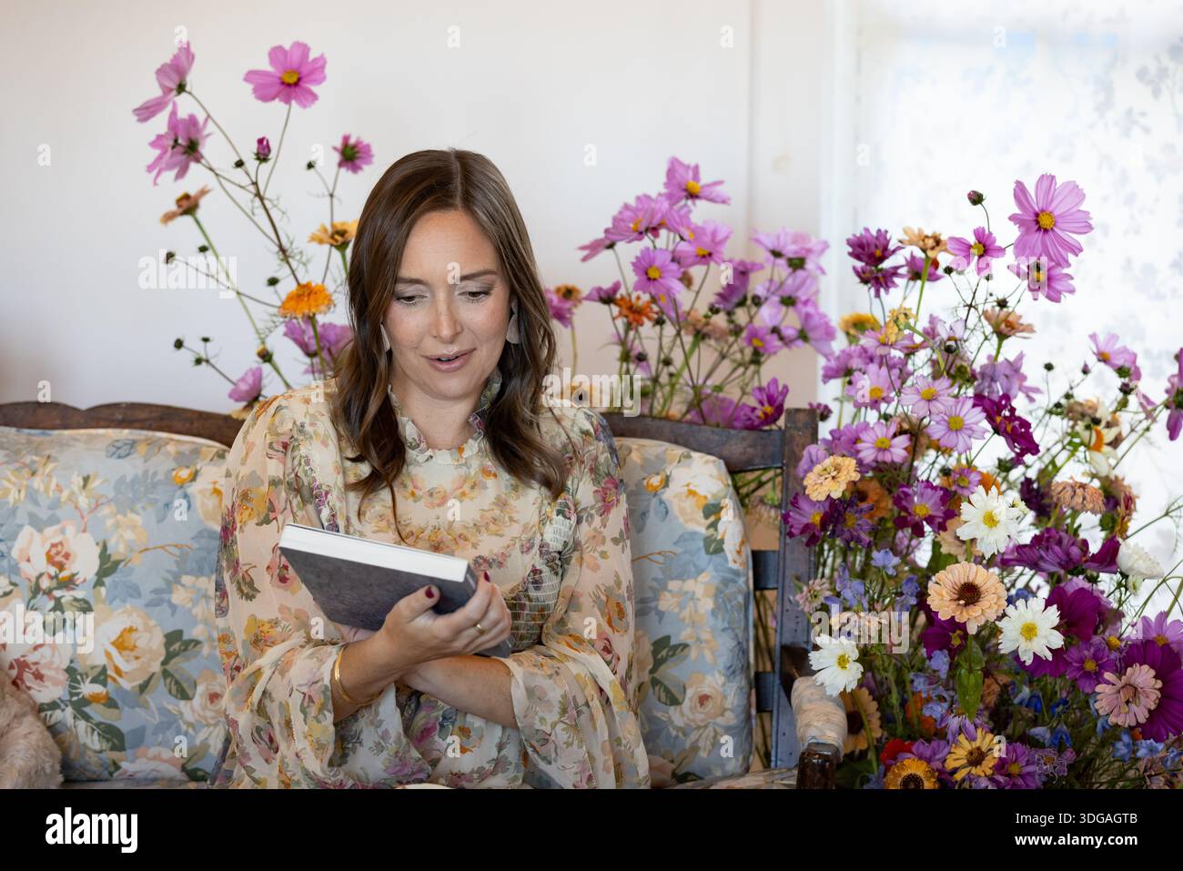 Eine Frau auf der Couch, umgeben von Blumen, schaut sich ein Buch an Stockfoto