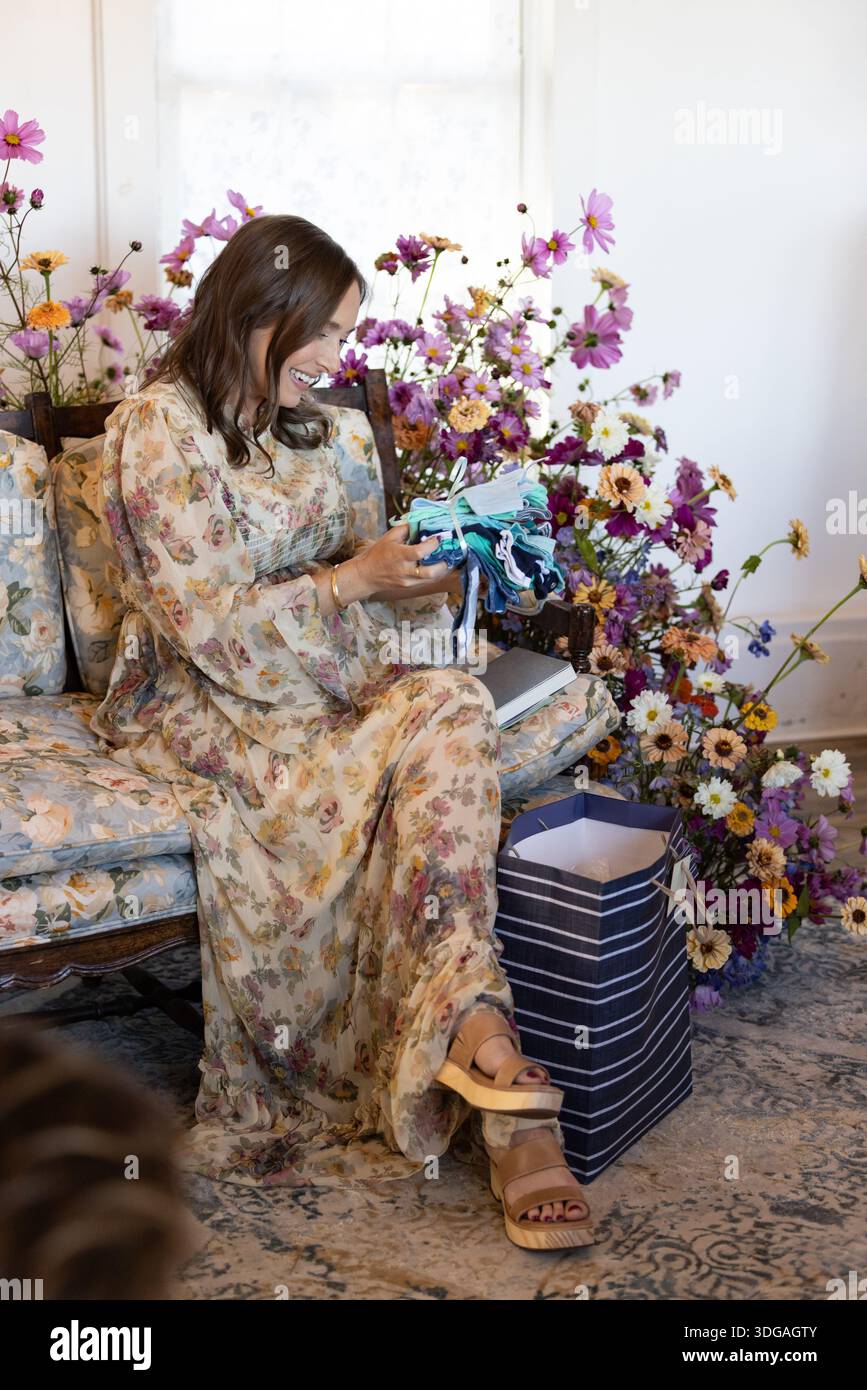 Frau auf Blumensofa packt Babyduschgeschenke aus, umgeben von Fl Stockfoto