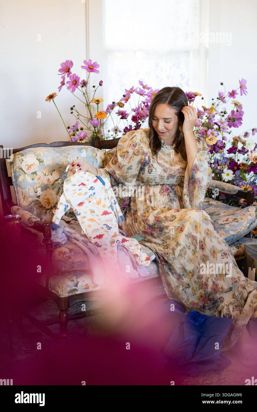 Frau auf Blumensofa packt Babyduschgeschenke aus, umgeben von Fl Stockfoto