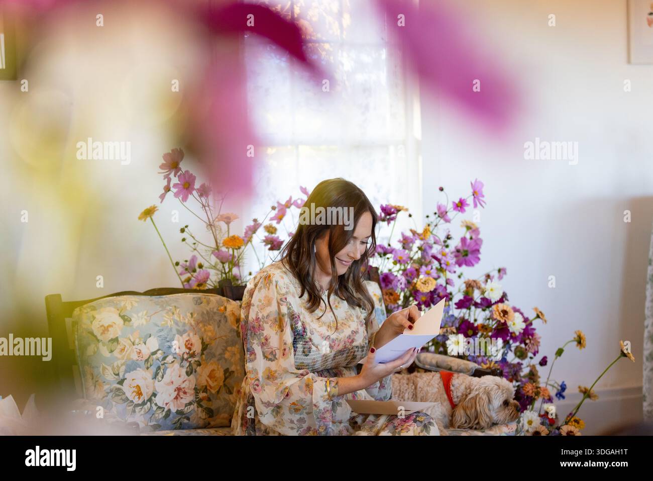 Frau auf Blumensofa, eingerahmt von Blumen, liest eine Karte Stockfoto