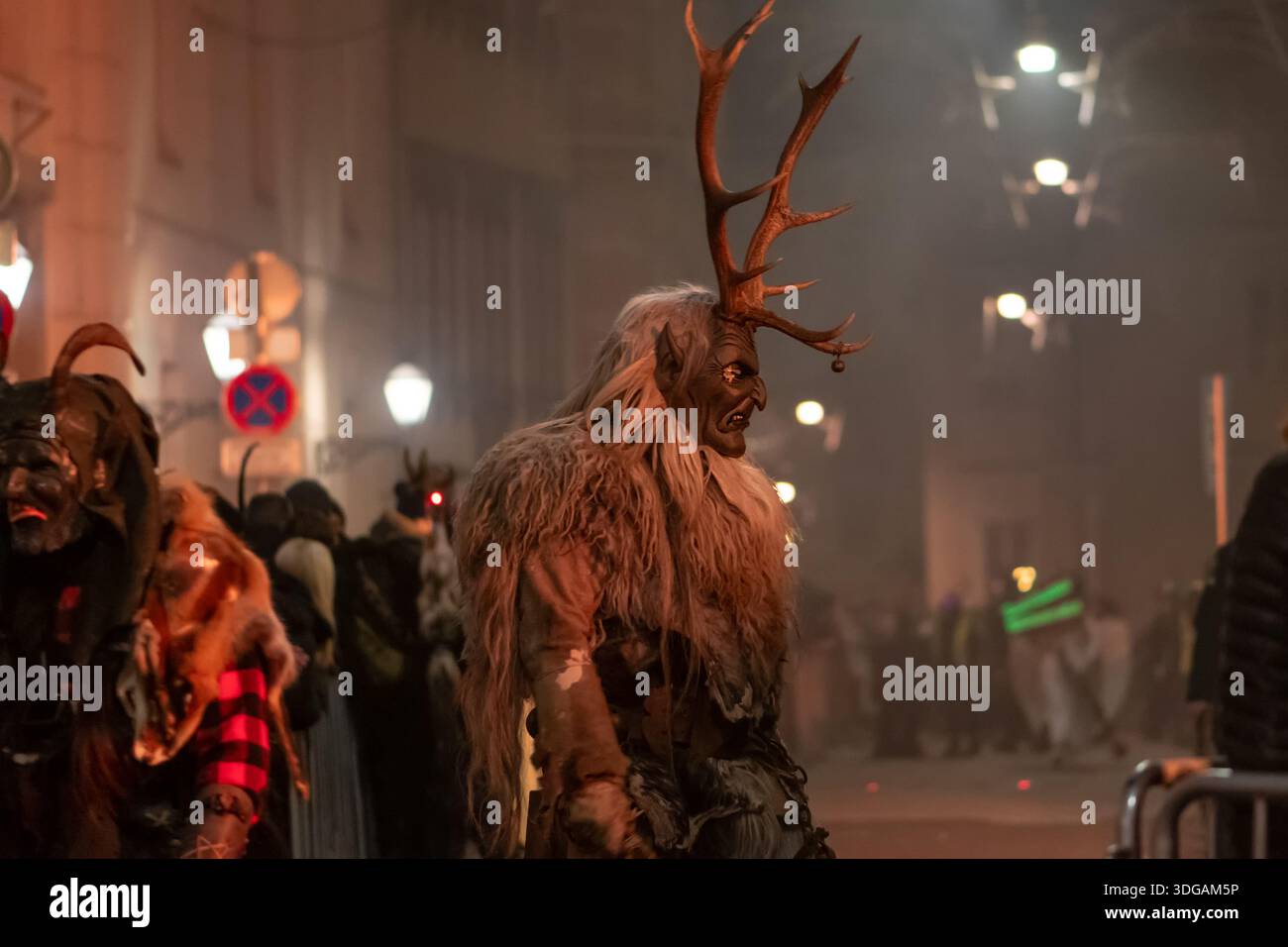 Krampus mit großen Hirschgeweihen spaziert neben anderen Prozessionsteilnehmern entlang der Straße Stockfoto
