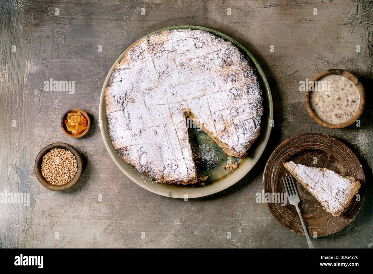 Hausgemachte Torte Pastiera Napolitano mit Puderzucker bestreut auf Keramikplatte mit Scheibe auf Holzplatte. Kleine Schüsseln mit Zutaten Stockfoto