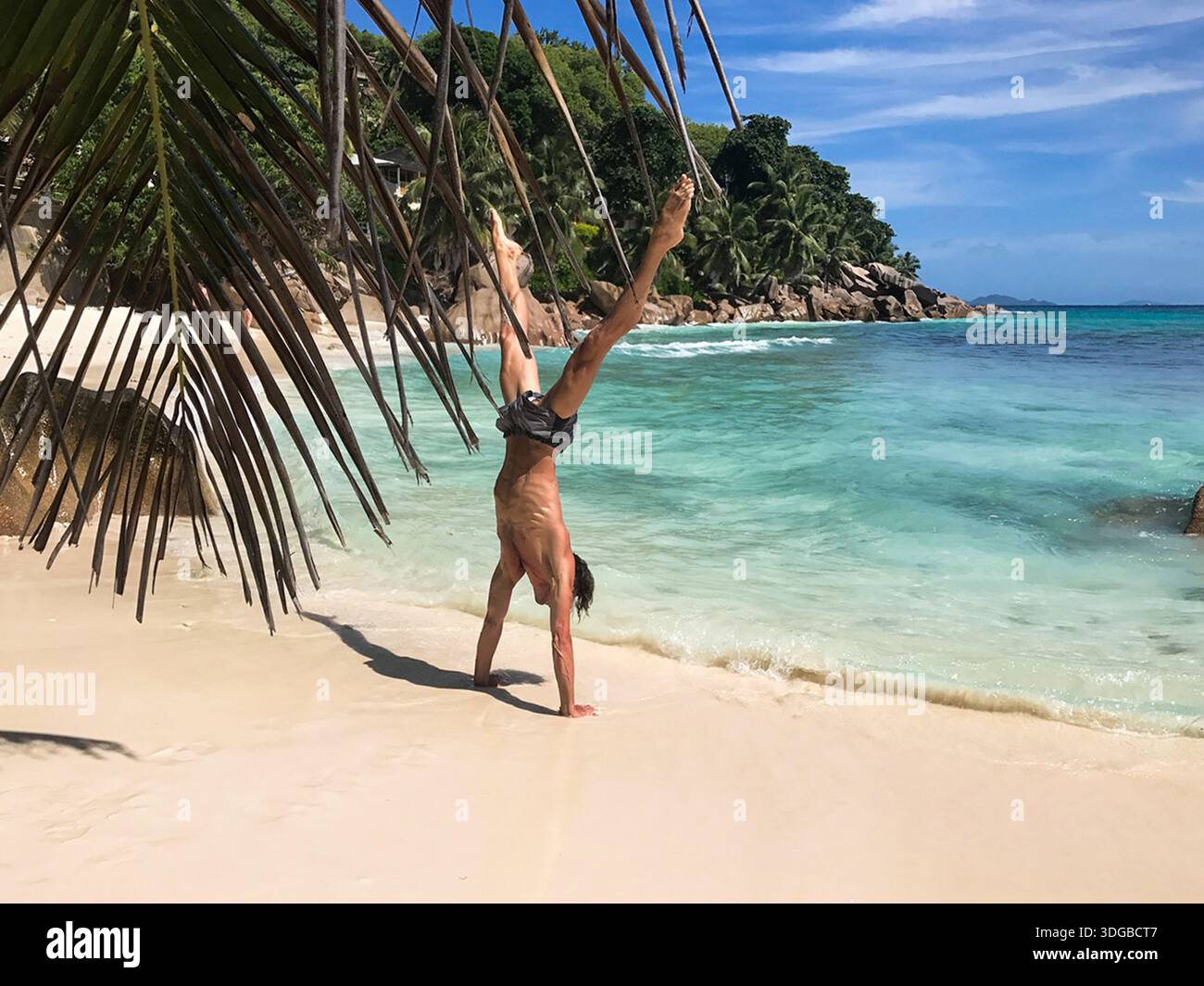 Sportlicher Mann, der einen Handstand mit auseinander liegenden Beinen an einem tropischen Strand mit weißem Sand und türkisfarbenem Wasser macht. Palmblätter umrahmen die Szene, mit grüner Küste. Stockfoto