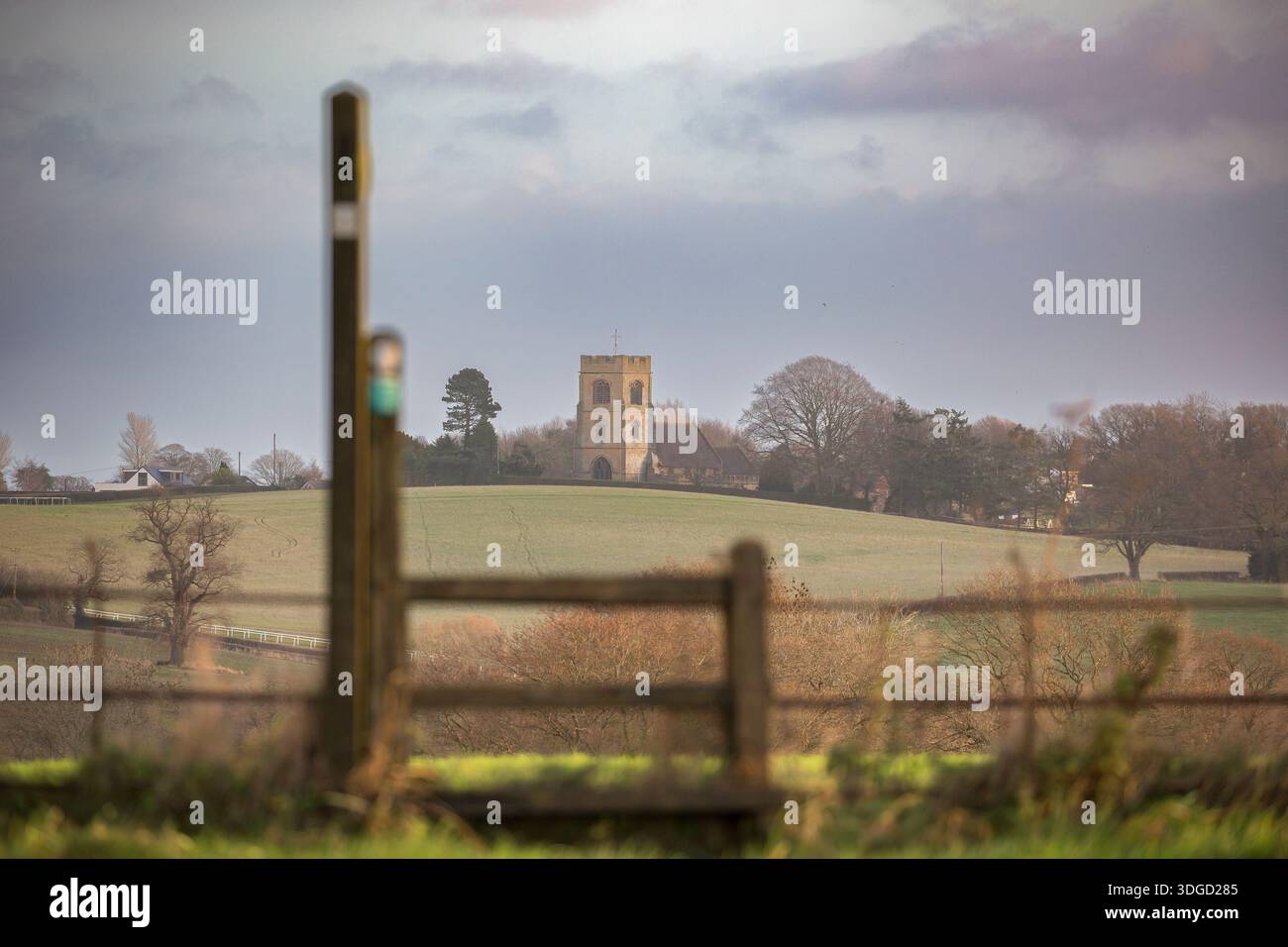 Fernansicht einer englischen Kirche über offene Landschaft und Felder mit einem Fußweg und einem Steg im Vordergrund. Stockfoto