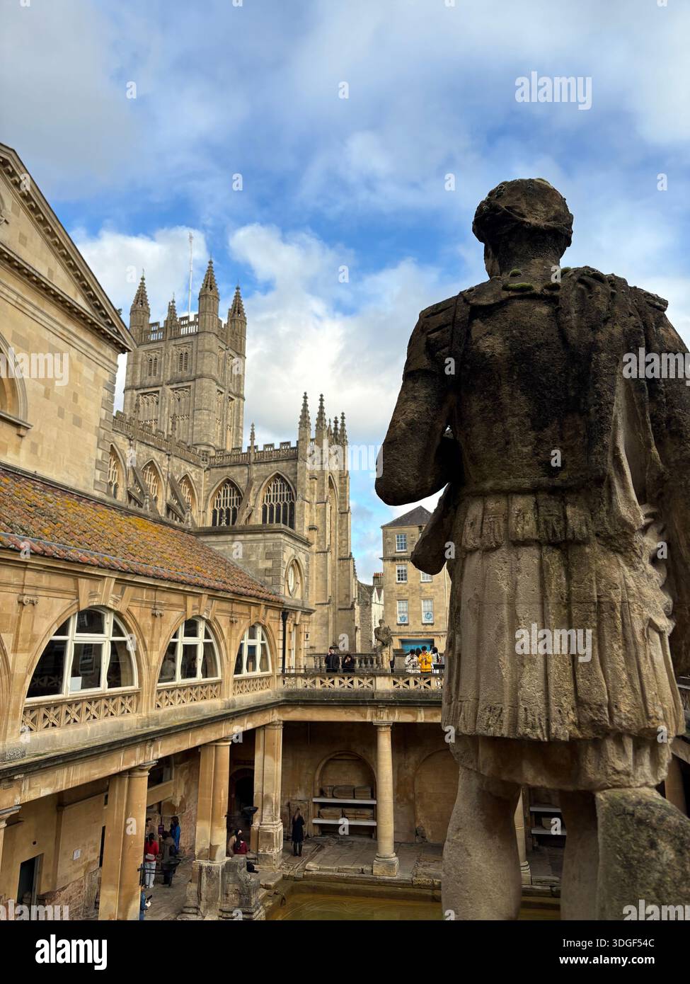 Historische Steinstatue mit Blick auf den Komplex der römischen Bäder mit Bath Abbey im Hintergrund in Bath, England, unter teilweise bewölktem Himmel. - Smartphone-aufgenommenes Stockfoto