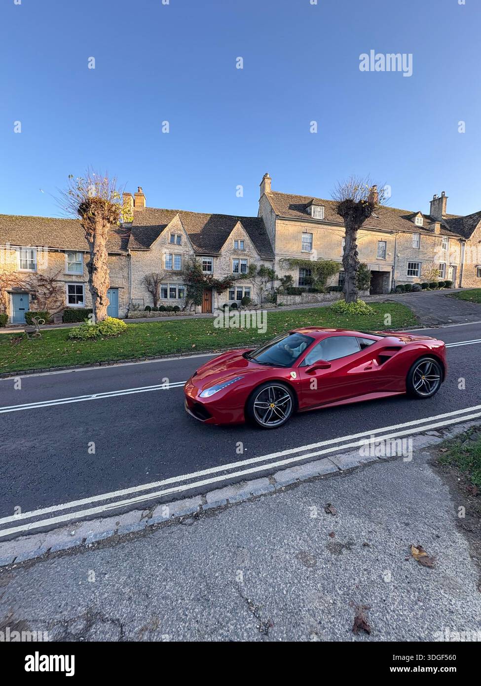 Roter Ferrari-Sportwagen, der durch eine traditionelle Dorfstraße von Cotswolds mit historischen Steinhäusern im Hintergrund fährt, England. - Smartphone-aufgenommenes Stockfoto