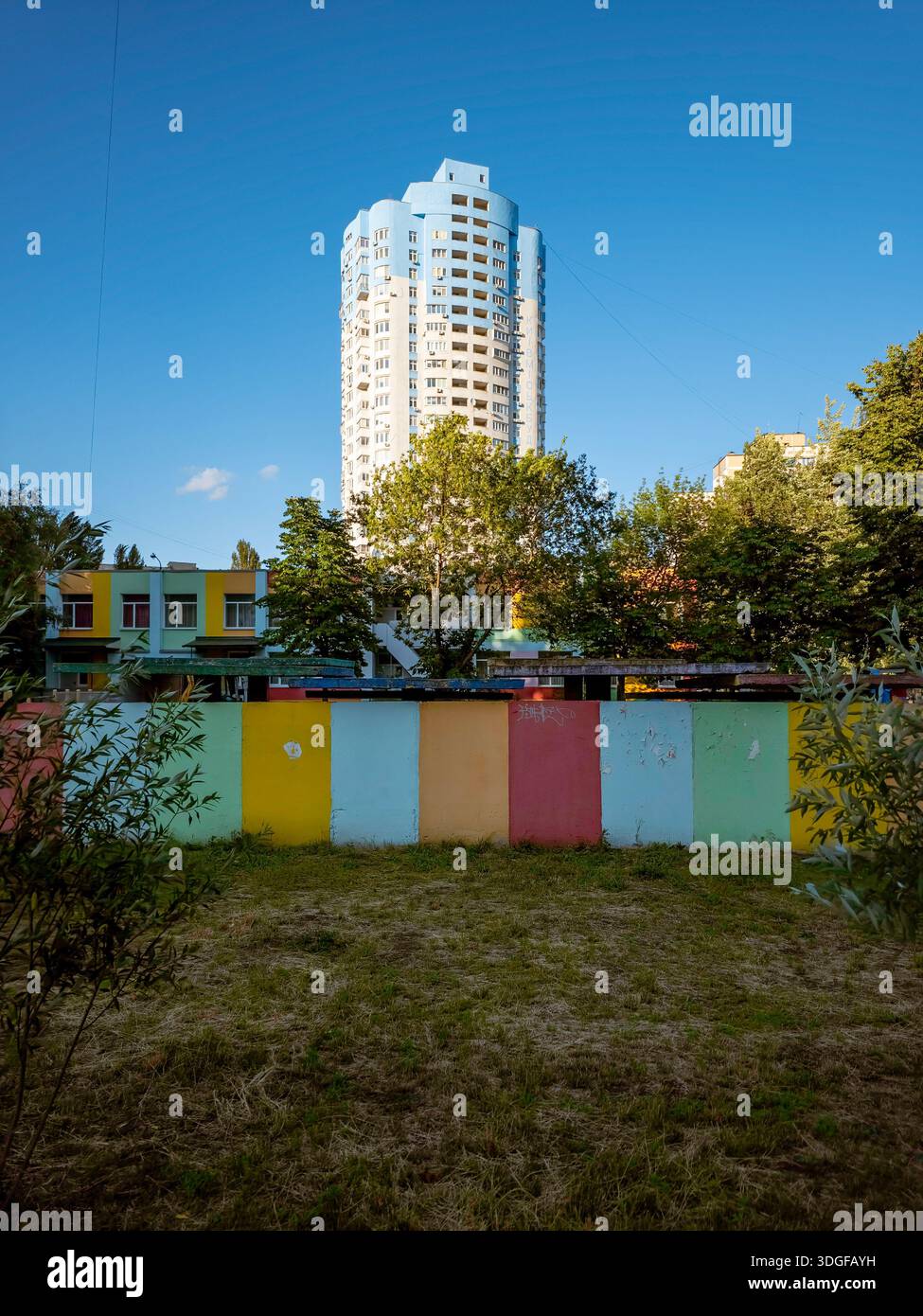 Kiew, Ukraine - 27. Juni 2025: Das Hochhaus in der Revutsky Street 7V erhebt sich hinter einem bunten Kindergartenzaun und dem Kindergartengebäude selbst, mit - Smartphone-aufgenommenes Stockfoto