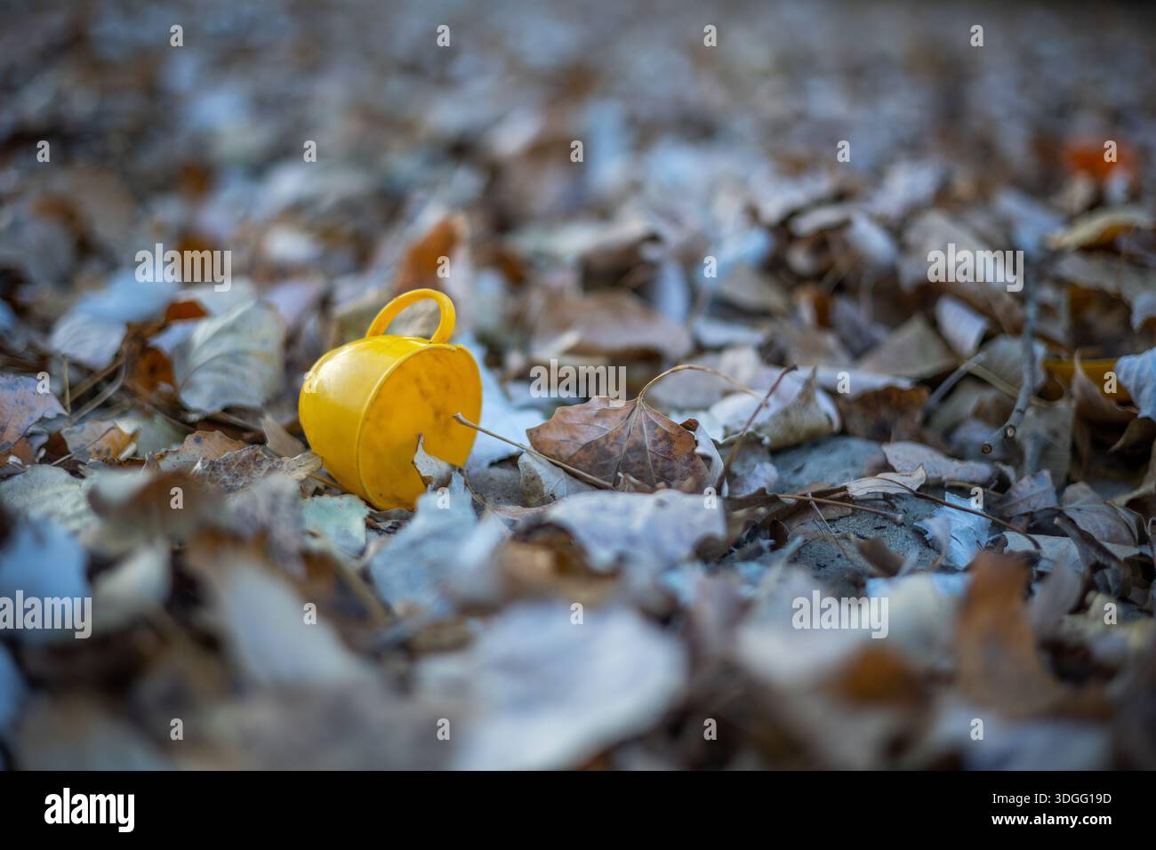 Ein kleiner gelber Spielzeugbecher liegt auf seiner Seite inmitten eines mit zahlreichen herbstlichen Pappelblättern bedeckten Bodens in Braun- und Beigetönen, unter Naturstein Stockfoto