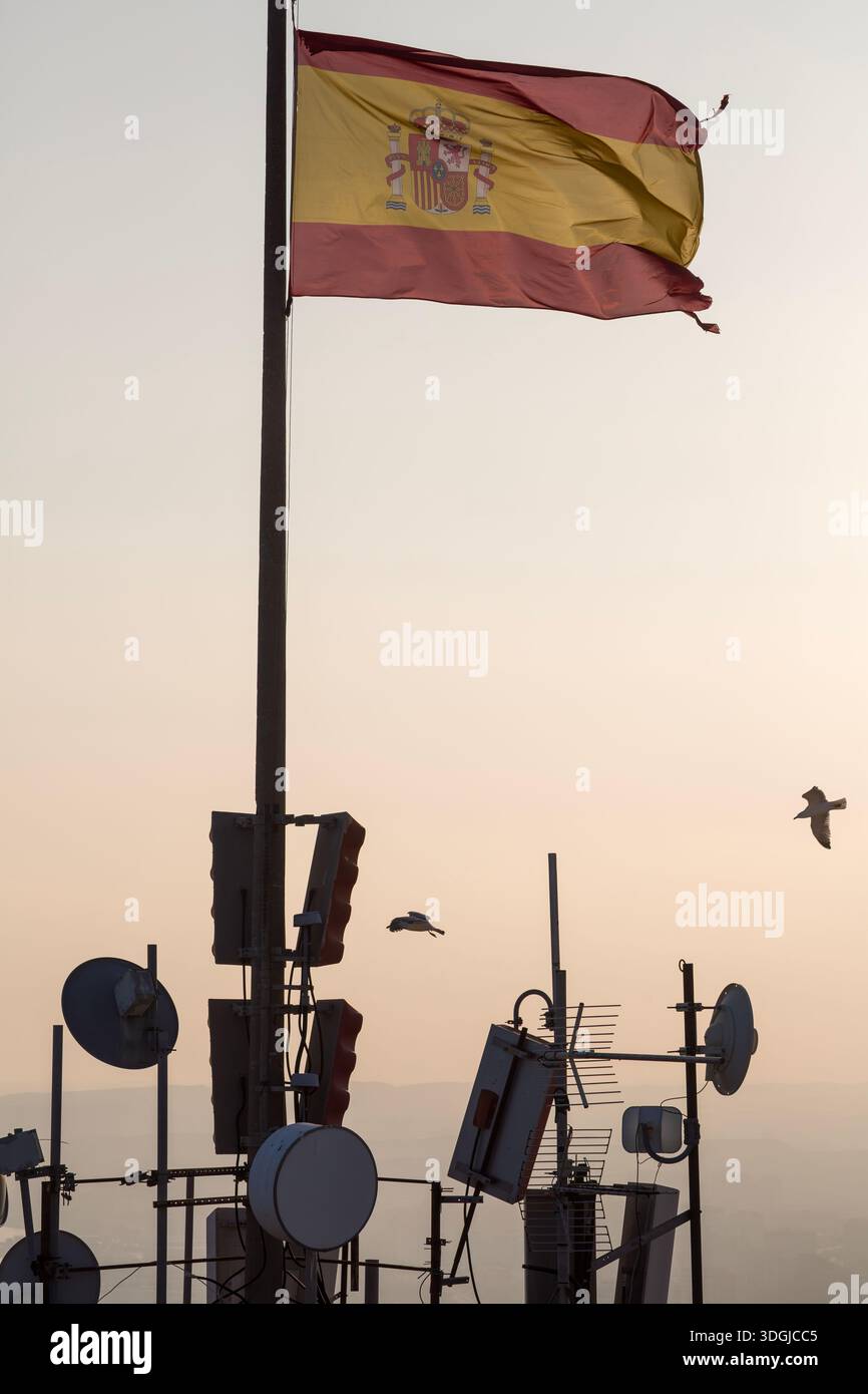Die Flagge Spaniens winkt von einem Telekommunikationsturm gegen einen warmen Sonnenuntergangshimmel, während Vögel vorbeifliegen. Ein Konzept der nationalen Kommunikationsinfrastruktur und der f Stockfoto