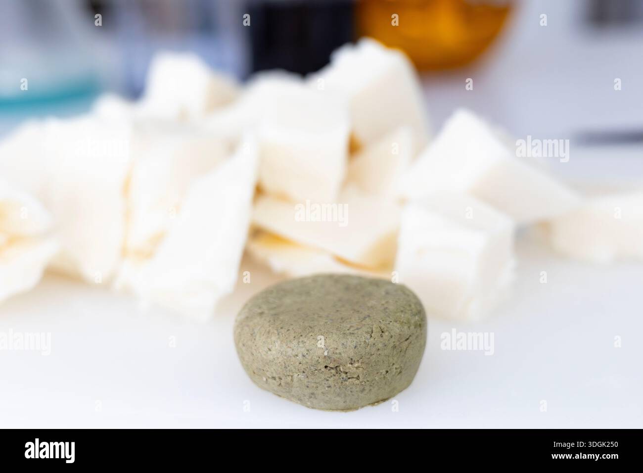 Eine runde, strukturierte, feste Shampoo-Bar sitzt auf einer weißen Oberfläche. Im verschwommenen Hintergrund befinden sich große Stücke roher Sheabutter, eine wichtige Zutat für Makin Stockfoto