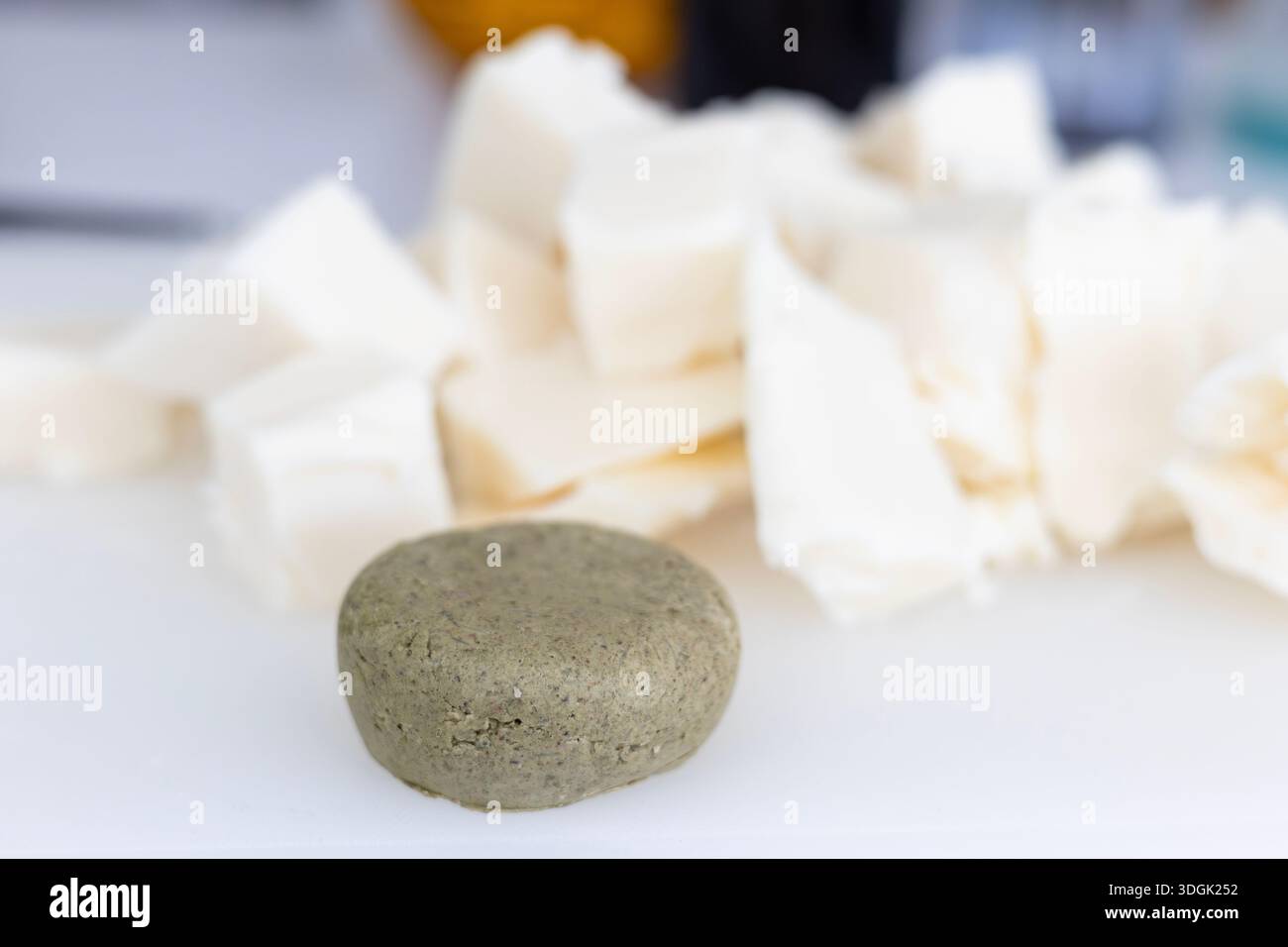 Eine runde, strukturierte, feste Shampoo-Bar sitzt auf einer weißen Oberfläche. Im verschwommenen Hintergrund befinden sich große Stücke roher Sheabutter, eine wichtige Zutat für Makin Stockfoto