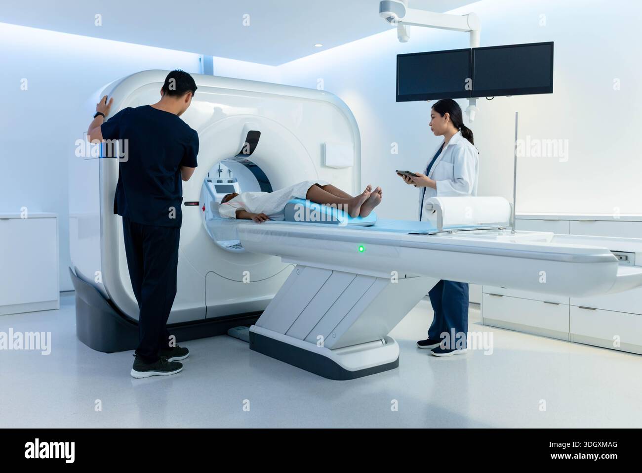 Diverse medizinische Teams, die weibliche Patientin im Kleid auf dem MRT-Tisch in der Scan-Suite betreuen, Tablette halten Stockfoto