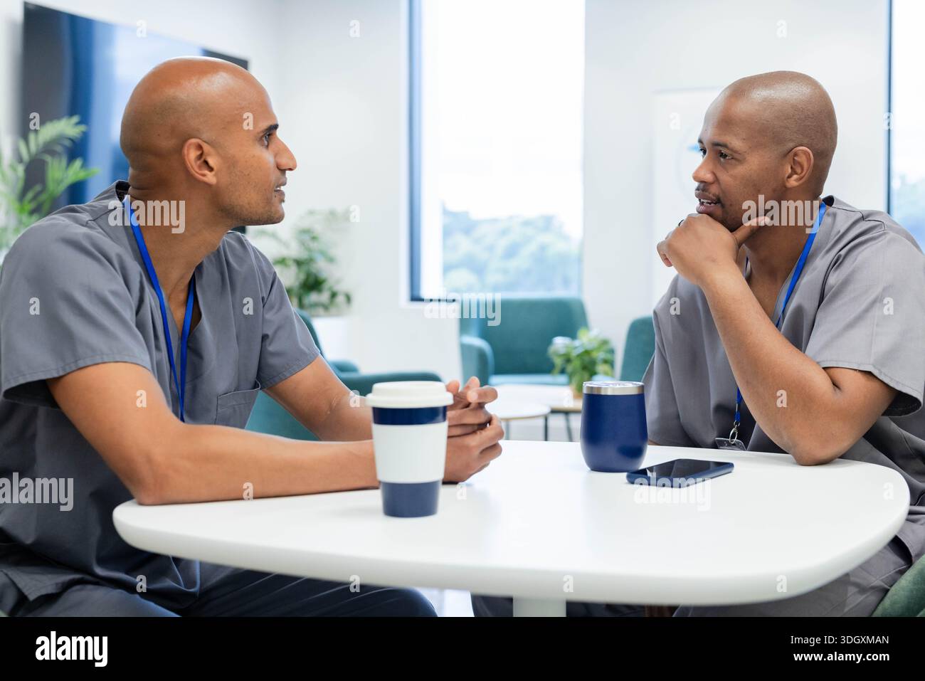 Afroamerikanische männliche Angestellte sitzen und sprechen in der Stabslounge in grauen Schutten, blauen Schlüsselbändern, Tassen Stockfoto